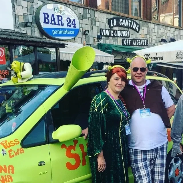 Two people standing next to a bright yellow vehicle with an oversized green horn on top, parked outside a building with signs for a bar and liquor store. The woman has red hair and the man wears sunglasses; both are smiling.
