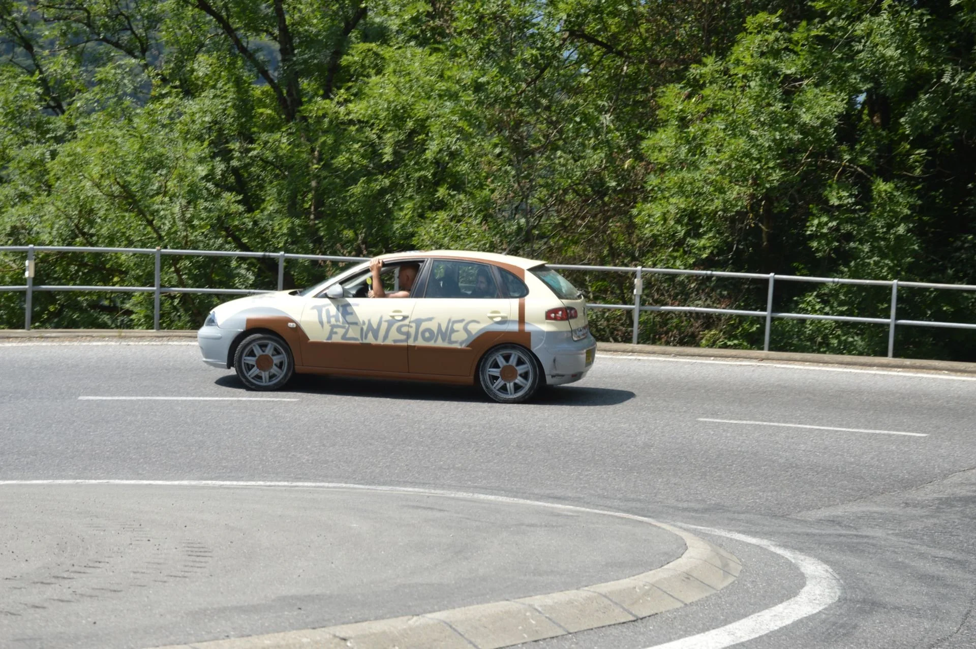 A small silver car with the words "The Flintstones" painted on its side, driving on a road with a guardrail and green trees in the background.