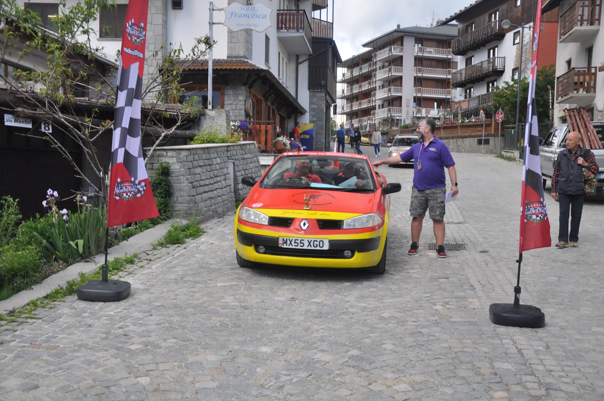 A yellow and red Renault car with a rally logo on the hood, parked on a cobblestone street. Two men are sitting inside the car, and another man is standing next to it, talking. There are two flags on stands flanking the car with racing symbols. The s