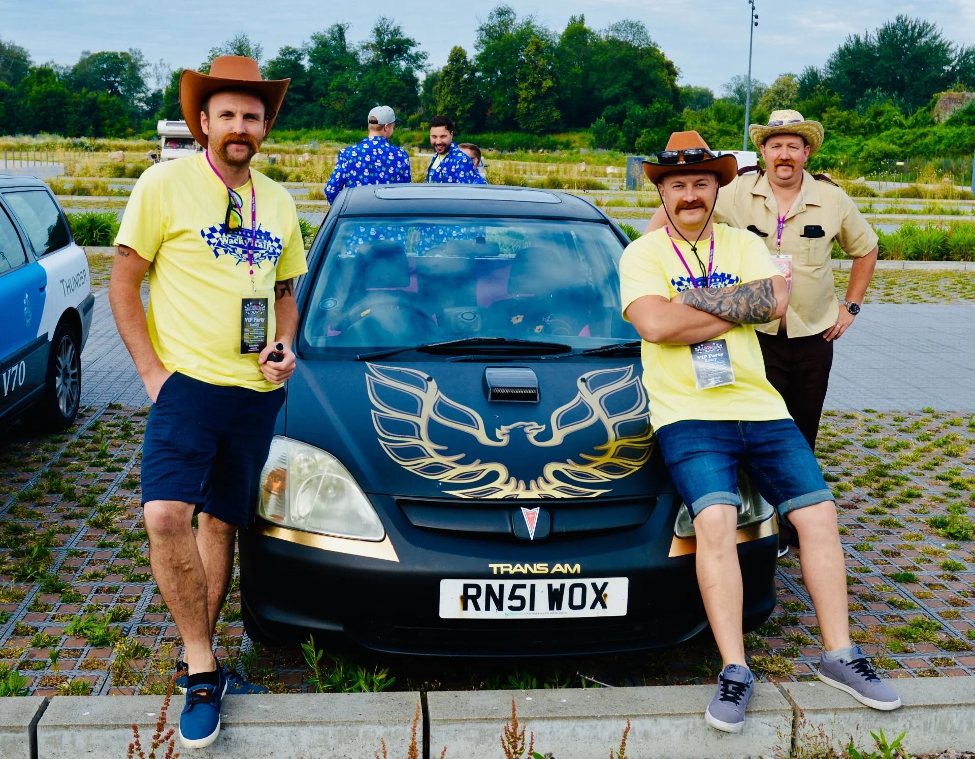 Four men standing around a black Pontiac Trans Am with a gold eagle decal on the hood, in an outdoor parking lot with trees in the background. Some men are wearing cowboy hats and yellow t-shirts, with one man leaning against the car with folded arms
