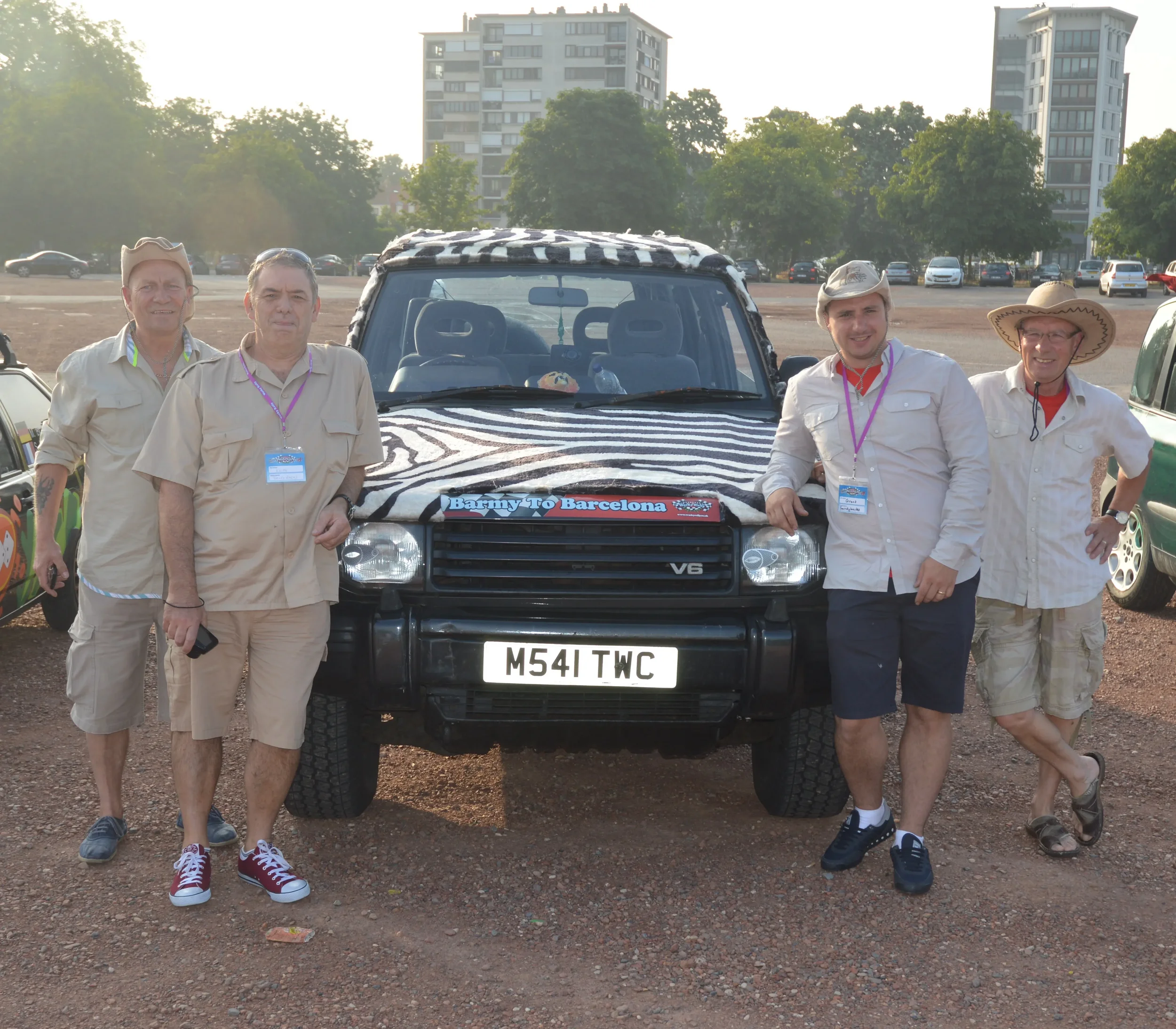 Five men in beige shirts and shorts standing next to a black vehicle with a zebra print cover and a 'Barney to Barcelona' sign, in a parking lot with trees and high-rise buildings in the background.