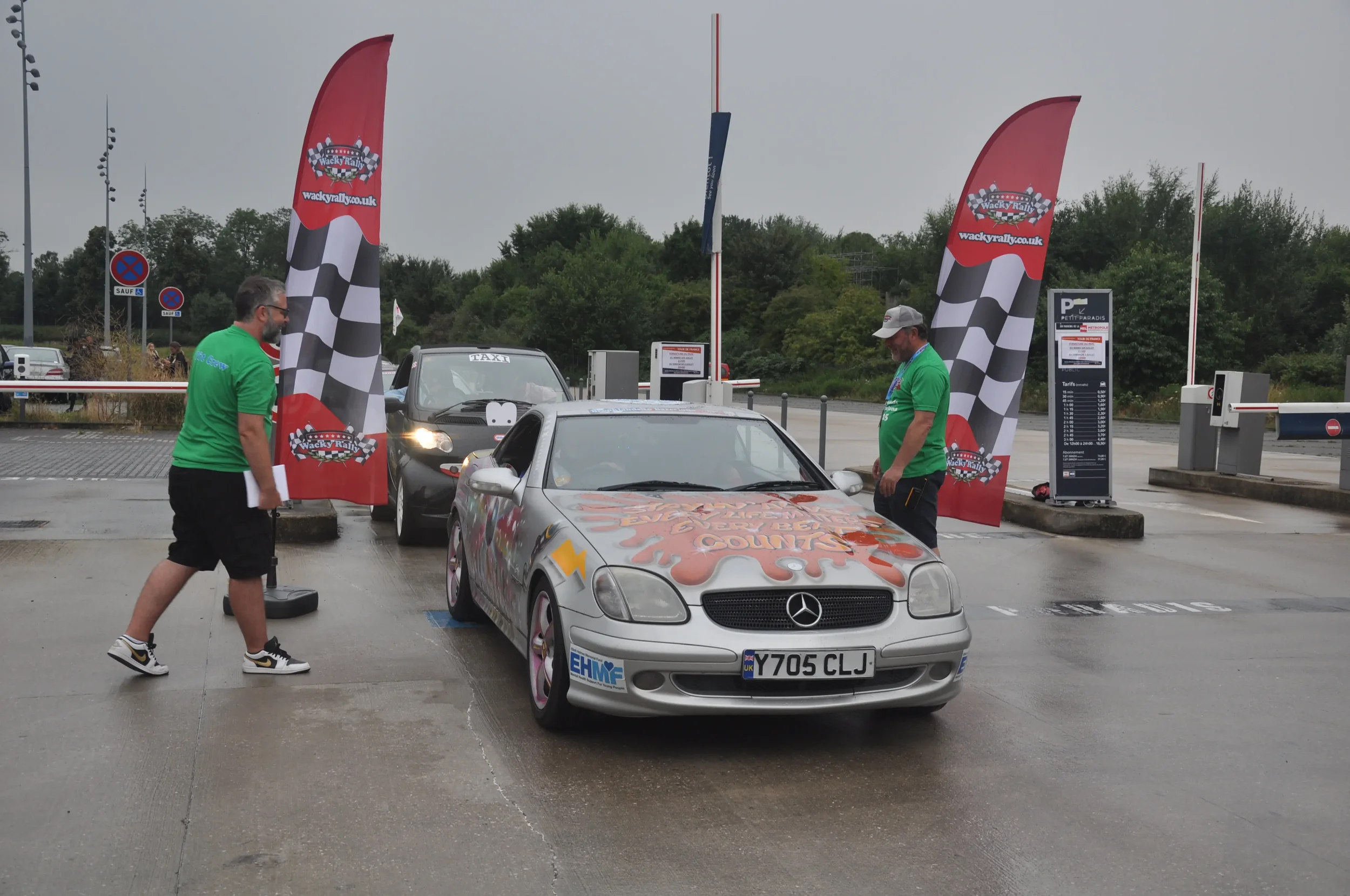 A silver Mercedes-Benz car at a racing event, with two men in green shirts standing nearby, one holding a clipboard. There are checkered flags, a parking lot, and a ticket booth in the background.