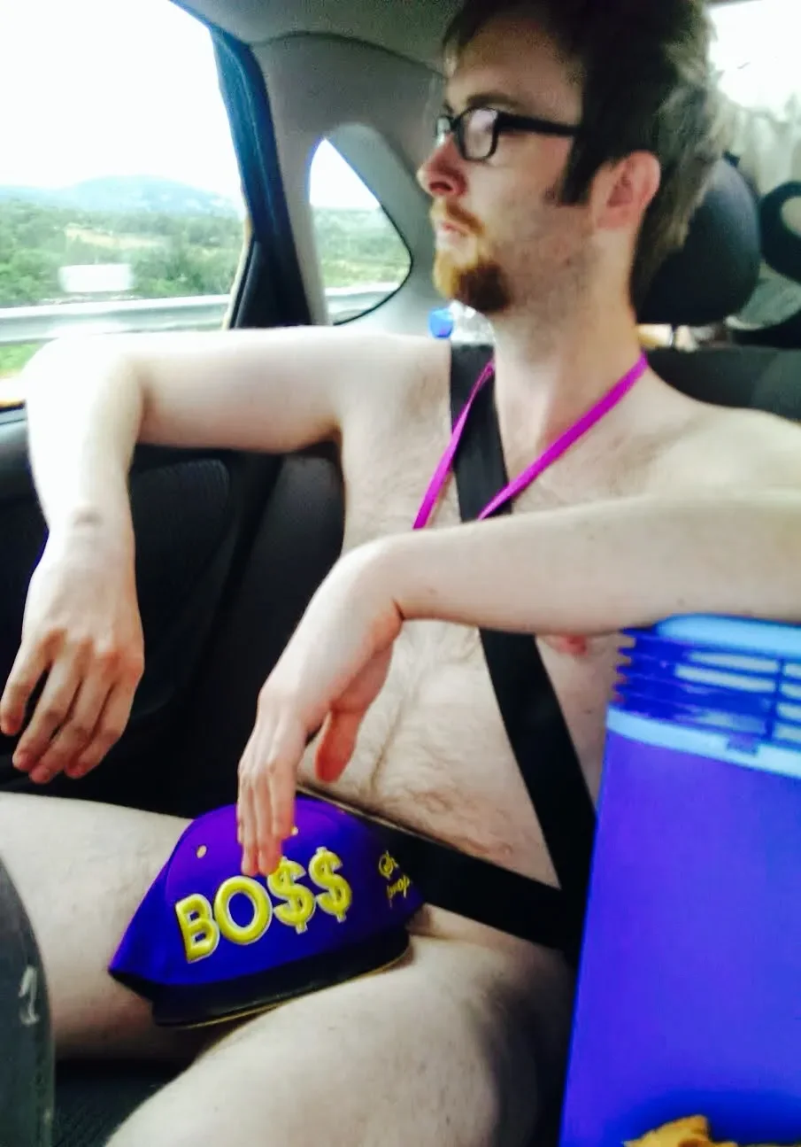 A shirtless young man with glasses and a beard sitting in the back seat of a car, holding party hats labeled 'BOSS$'. The man is relaxed, with his arms resting on the seat, and is looking out the window at a landscape with trees and hills.