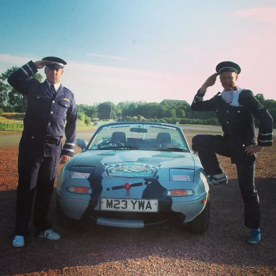 Two people in police officer costumes, one saluting the camera and the other with one foot on the car, standing next to a blue police-themed sports car outdoors on a sunny day.