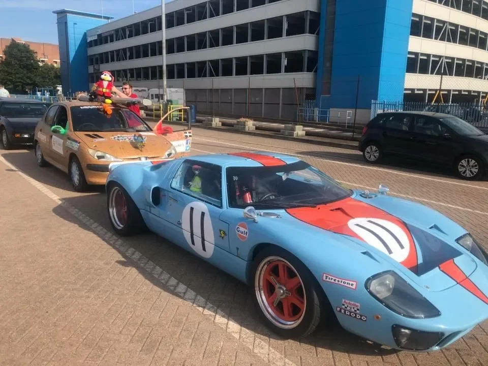 A blue vintage race car with red accents and racing numbers parked in a parking lot, with a tan car decorated for a parade behind it, featuring a person holding a stuffed animal on its roof.