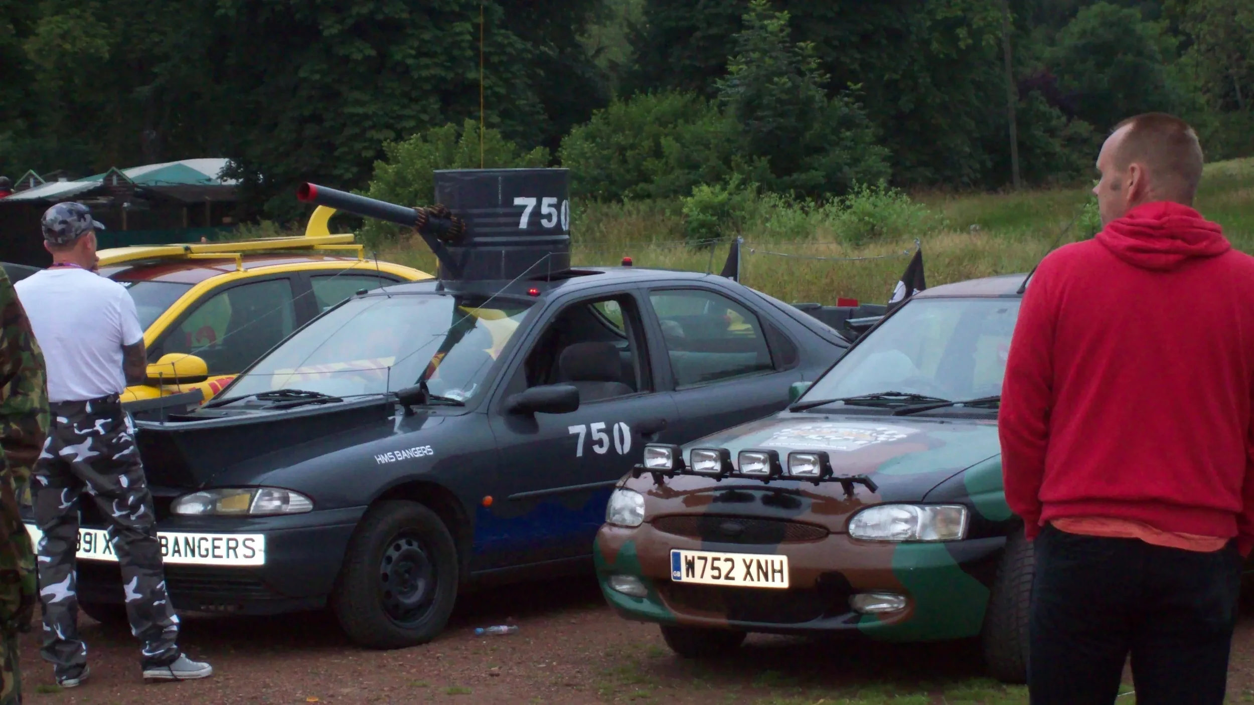 A black vehicle with military markings and the number 750 on it, parked next to a car with camouflage paint. Several people are standing around, including a person in a red hoodie and another in camouflage pants, with trees in the background.