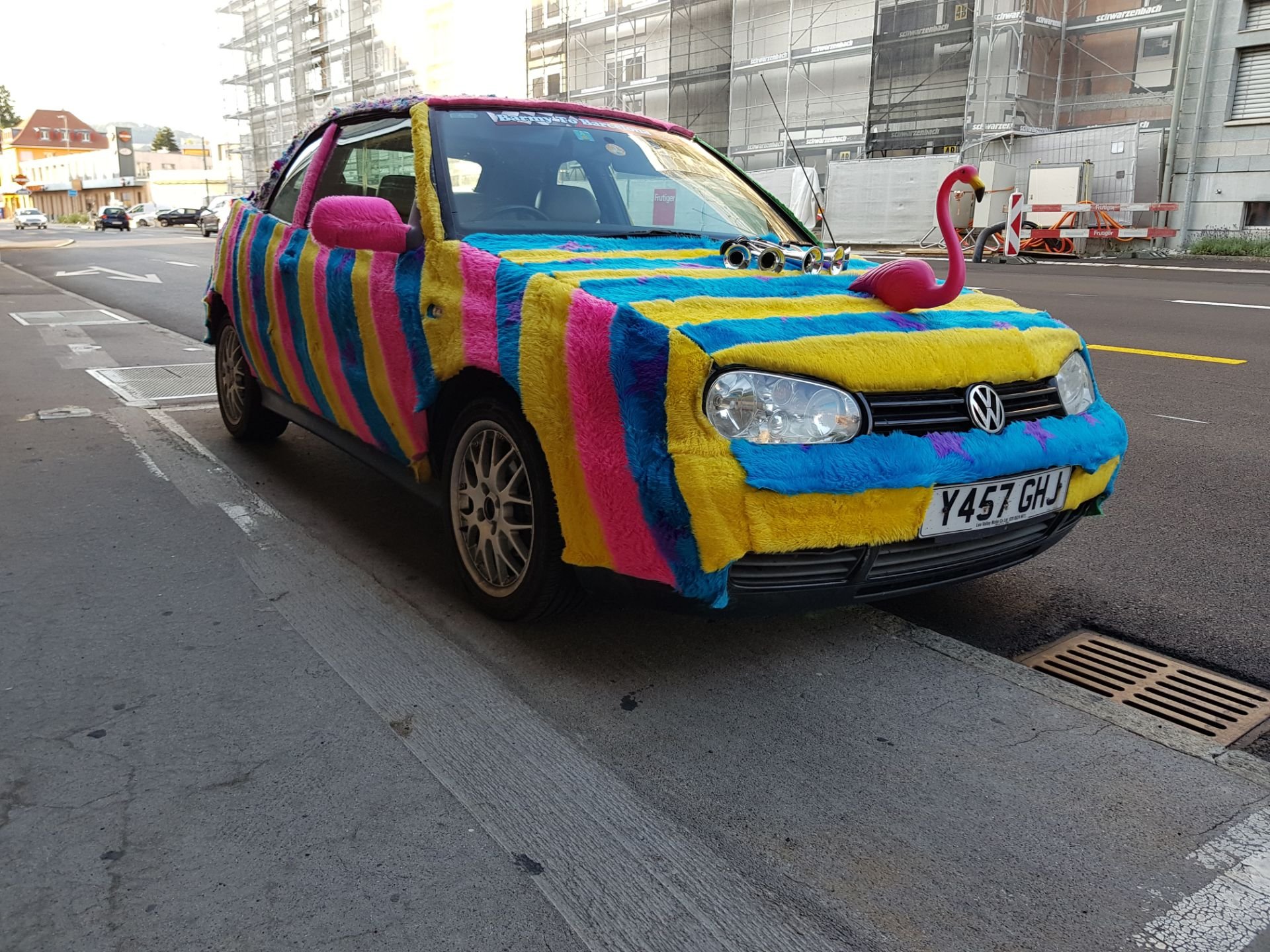 A Volkswagen car decorated with colorful fluffy fabric in pink, yellow, blue, and purple. A pink plastic flamingo is on the hood, and the car has multiple goggles on the roof.