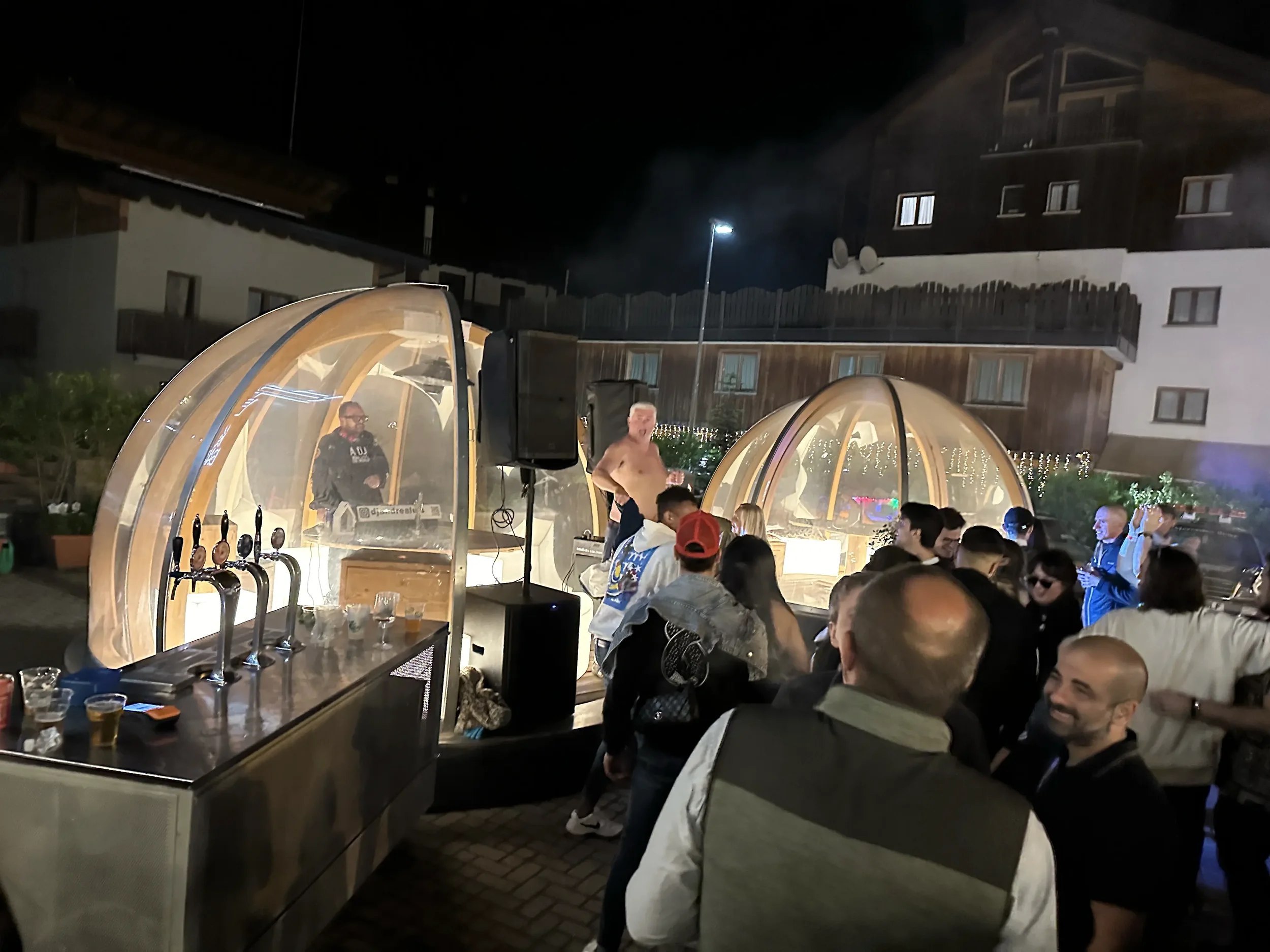 A nighttime outdoor party with people dancing and socializing under transparent dome-shaped tents. A DJ is playing music inside one dome, with some men shirtless and others wearing jackets. The scene includes tables with drinks and a bar area with be
