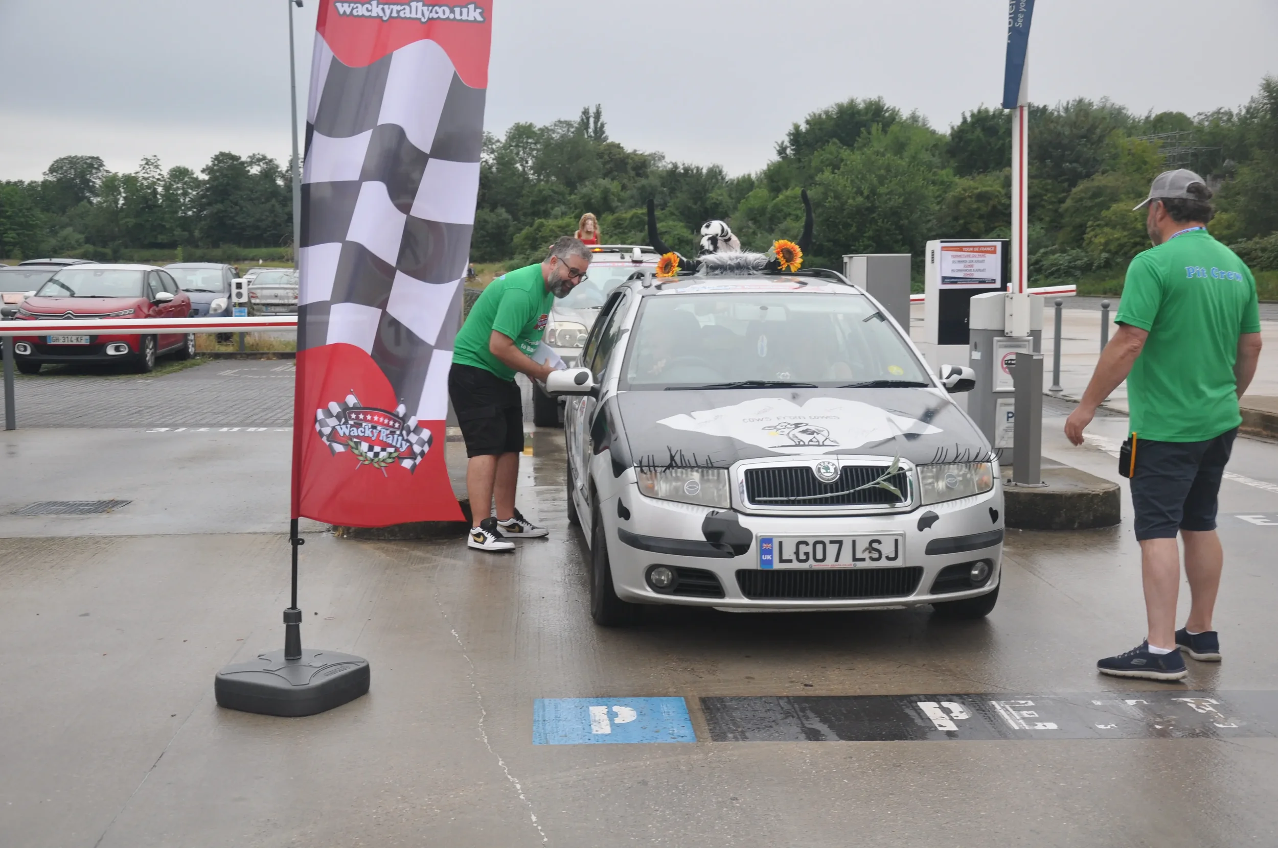 A silver car with a dog on the roof decorated with sunflowers and a cartoon drawing on the hood, parked in a parking lot during a racing event. Two men wearing green shirts stand beside the car, one looking at the car and the other standing further a