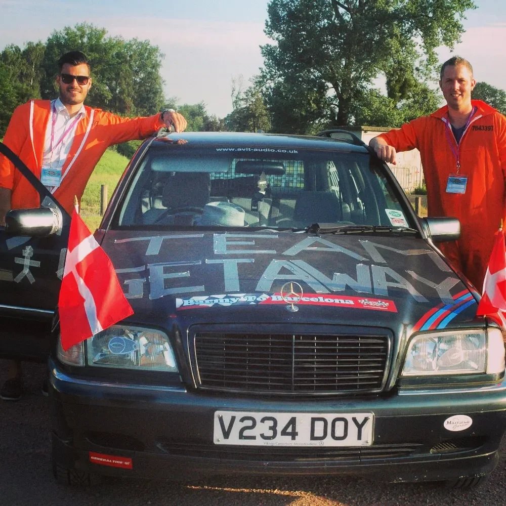 Two men in orange jackets standing beside a decorated black car with front flags, parked outdoors on a sunny day with green trees in the background.