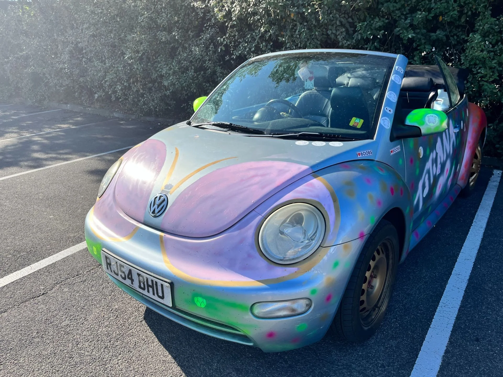 A Volkswagen Beetle convertible with rainbow and pastel-colored graffiti-style paint, parked in a marked parking space near bushes.