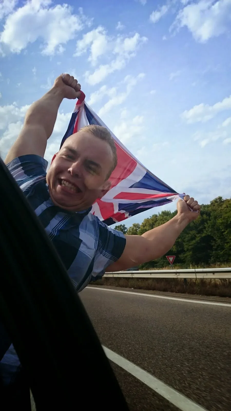 A man holding and waving a Union Jack flag while leaning out of a vehicle on a highway, with a blue sky and trees in the background.