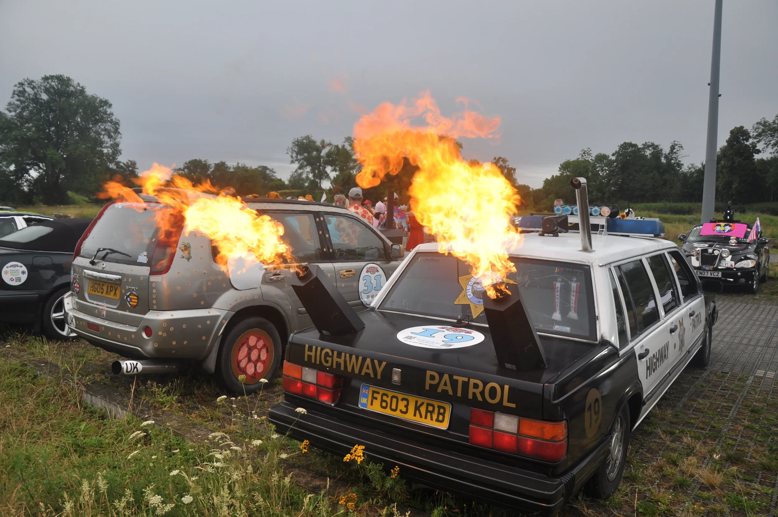 Several cars, including a highway patrol vehicle, are on fire with flames coming out of the hoods and rear of the vehicles, in an outdoor parking lot with trees and other parked cars in the background.