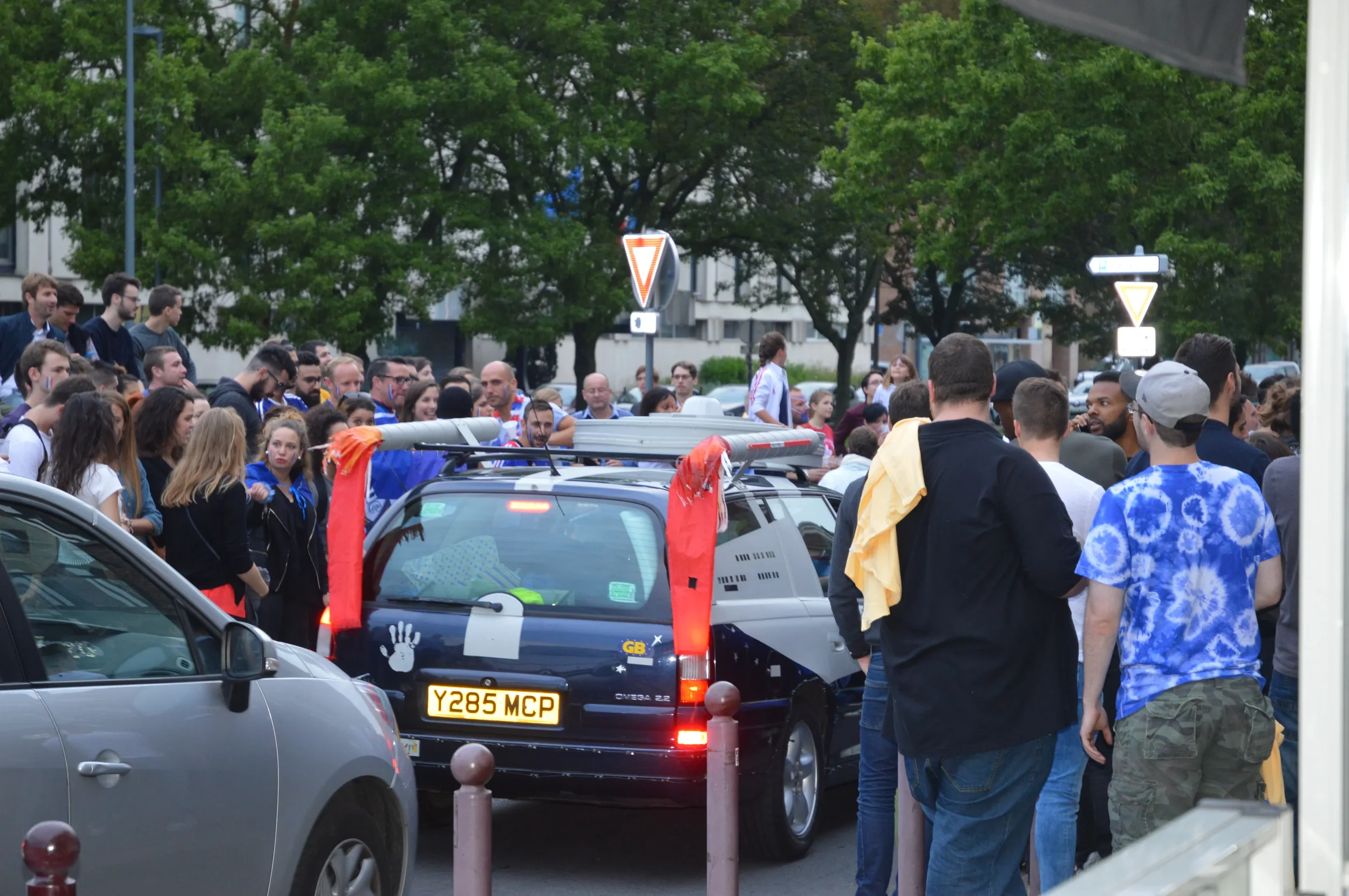 Crowd of people gathered outdoors around a black vehicle with a roof rack, some wearing casual clothing, and a few with yellow and red cloths tied to the vehicle, in front of green trees and street signs.