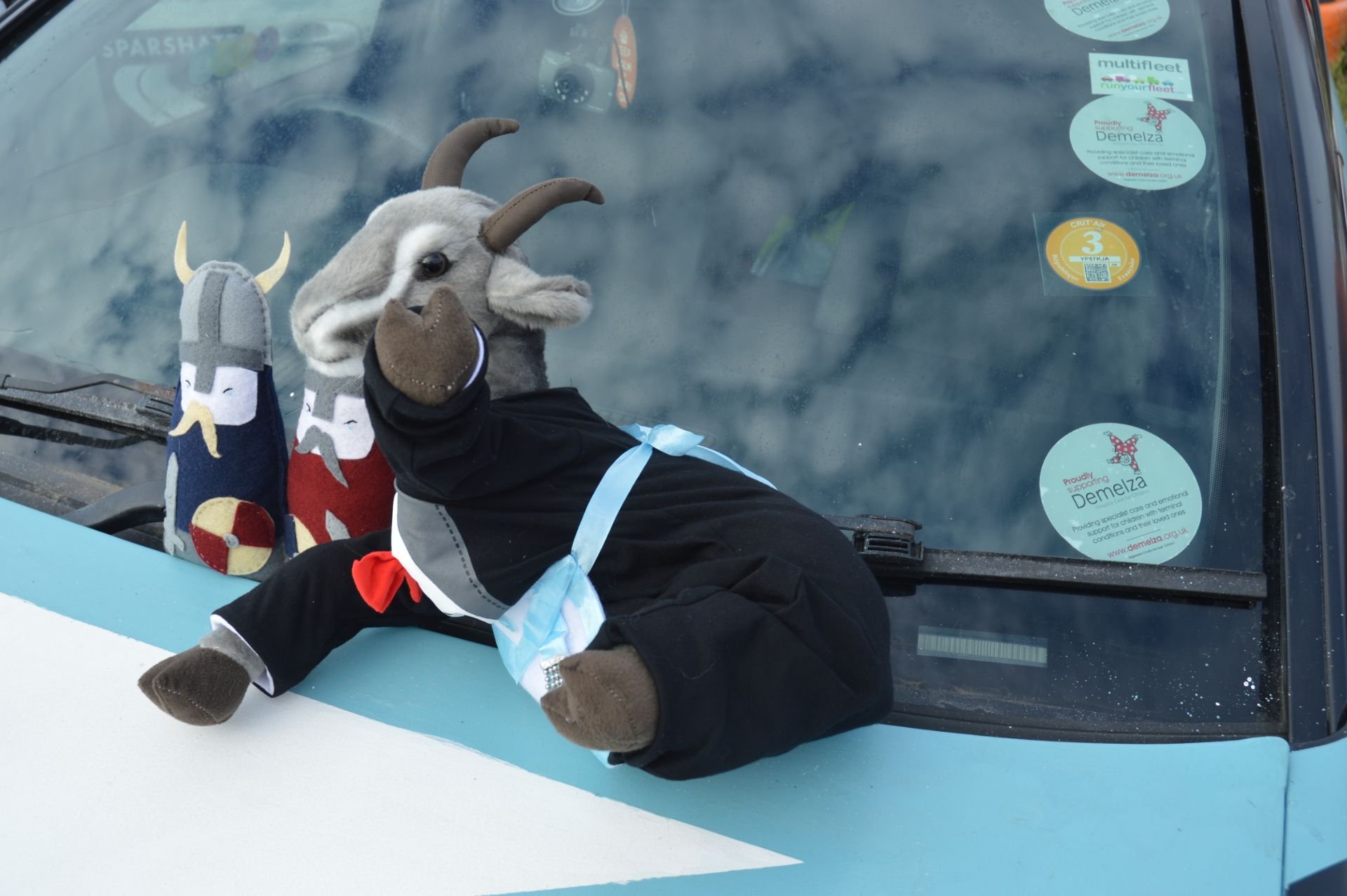 Stuffed goat and sock puppets displaying on the dashboard of a vehicle, with stickers on the windshield, including one for Demelza.
