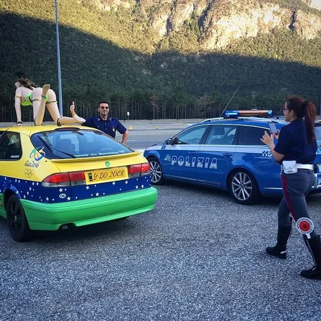 A man is standing next to a yellow and green car with an Olympic logo on it, giving a thumbs up. A police officer with a radio is standing beside a police car, both vehicles are on the side of a road with a mountainous background.