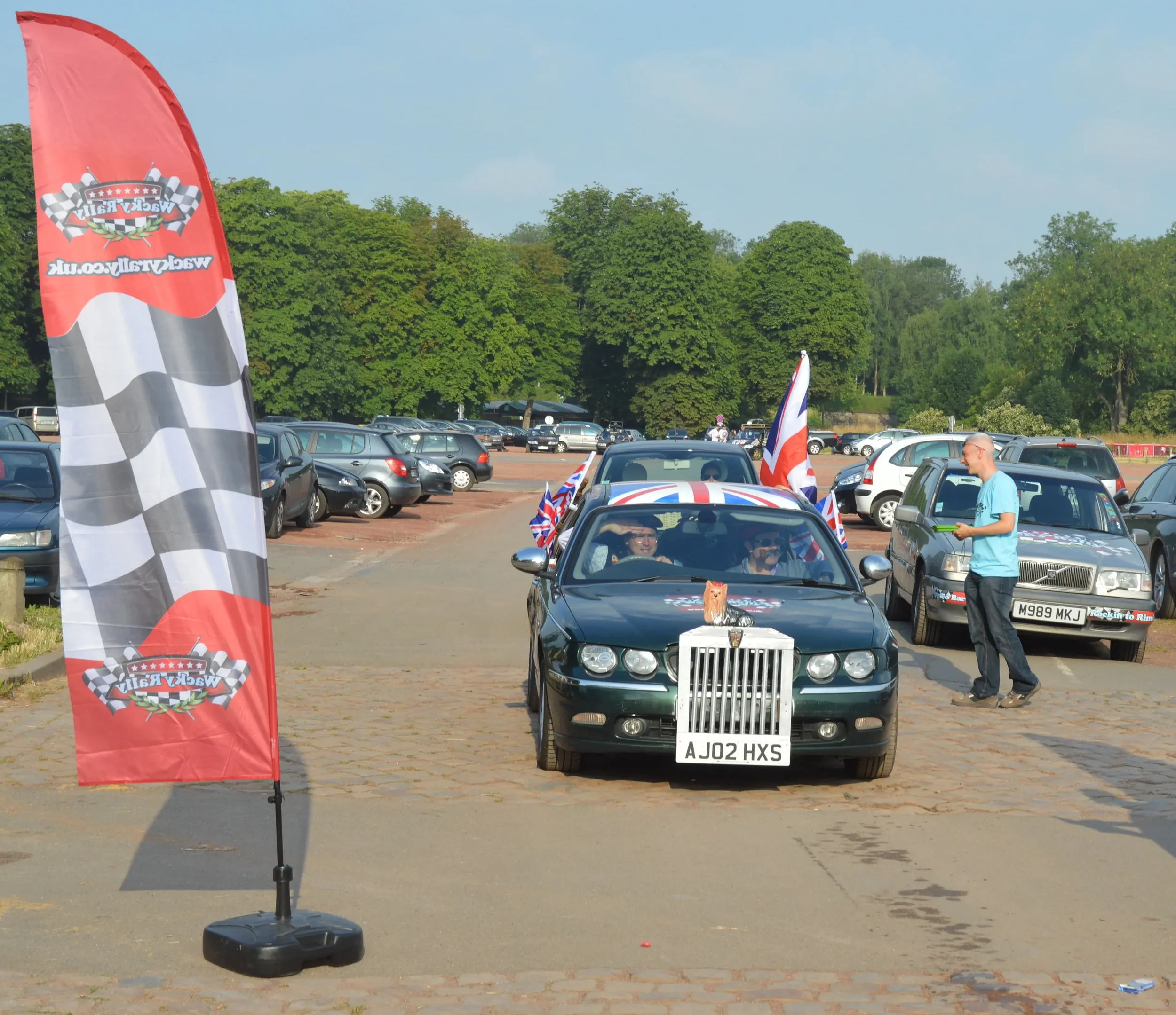 A black car decorated with British flags and a Union Jack roof, parked in a lot with a racing checkered flag banner on the left. There is a small stuffed animal on the front of the car, and people are seated inside. A person is standing next to the c