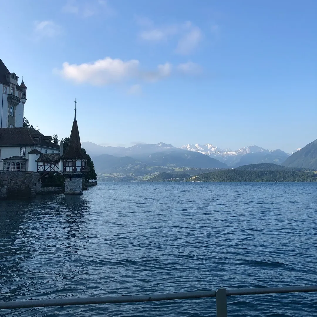 A scenic view of a large lake with mountains in the background, partly snow-capped, and a castle-like building on the left side of the image.