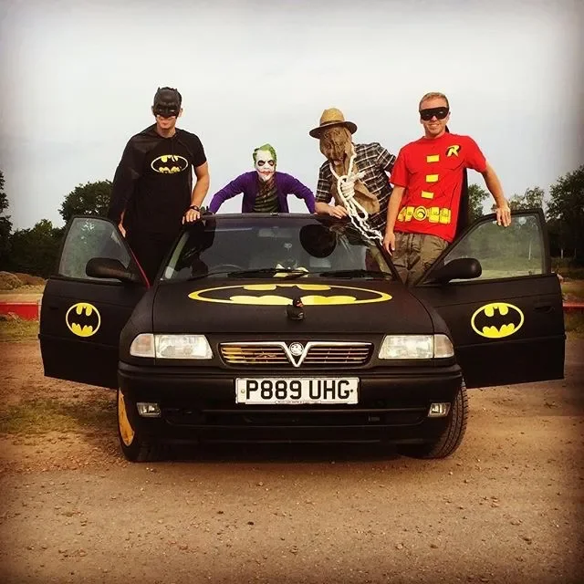 Four people dressed as Batman, Joker, and Robin stand around a black car decorated with Batman logos, with open doors and a license plate P889 UHG, on a dirt field with trees in the background.