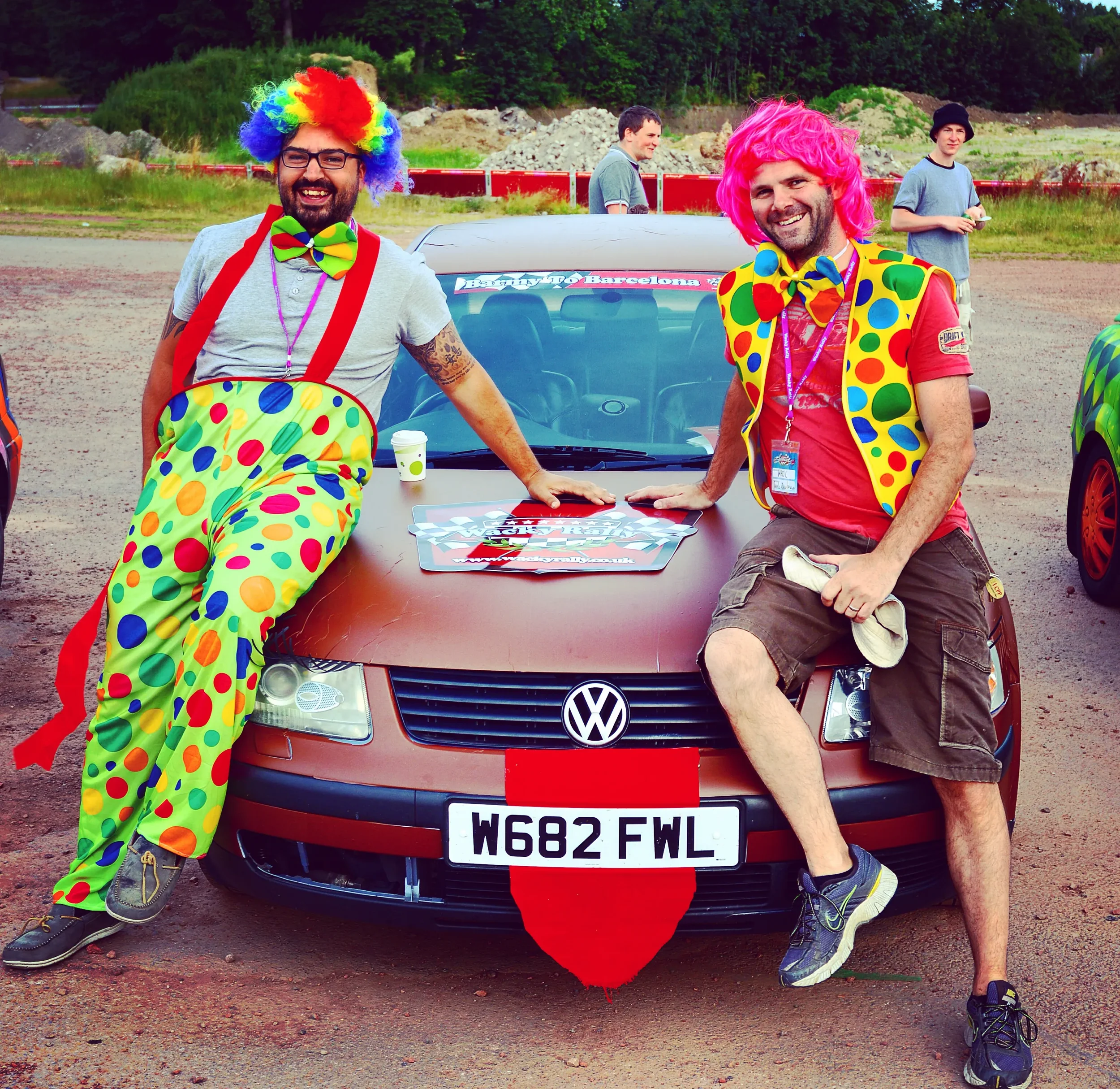 Two men dressed as clowns in colorful outfits are leaning against the front of a brown Volkswagen car, smiling at the camera. One is wearing a rainbow wig, large glasses, a bow tie, and polka-dotted pants, while the other has a pink wig, a polka-dot 