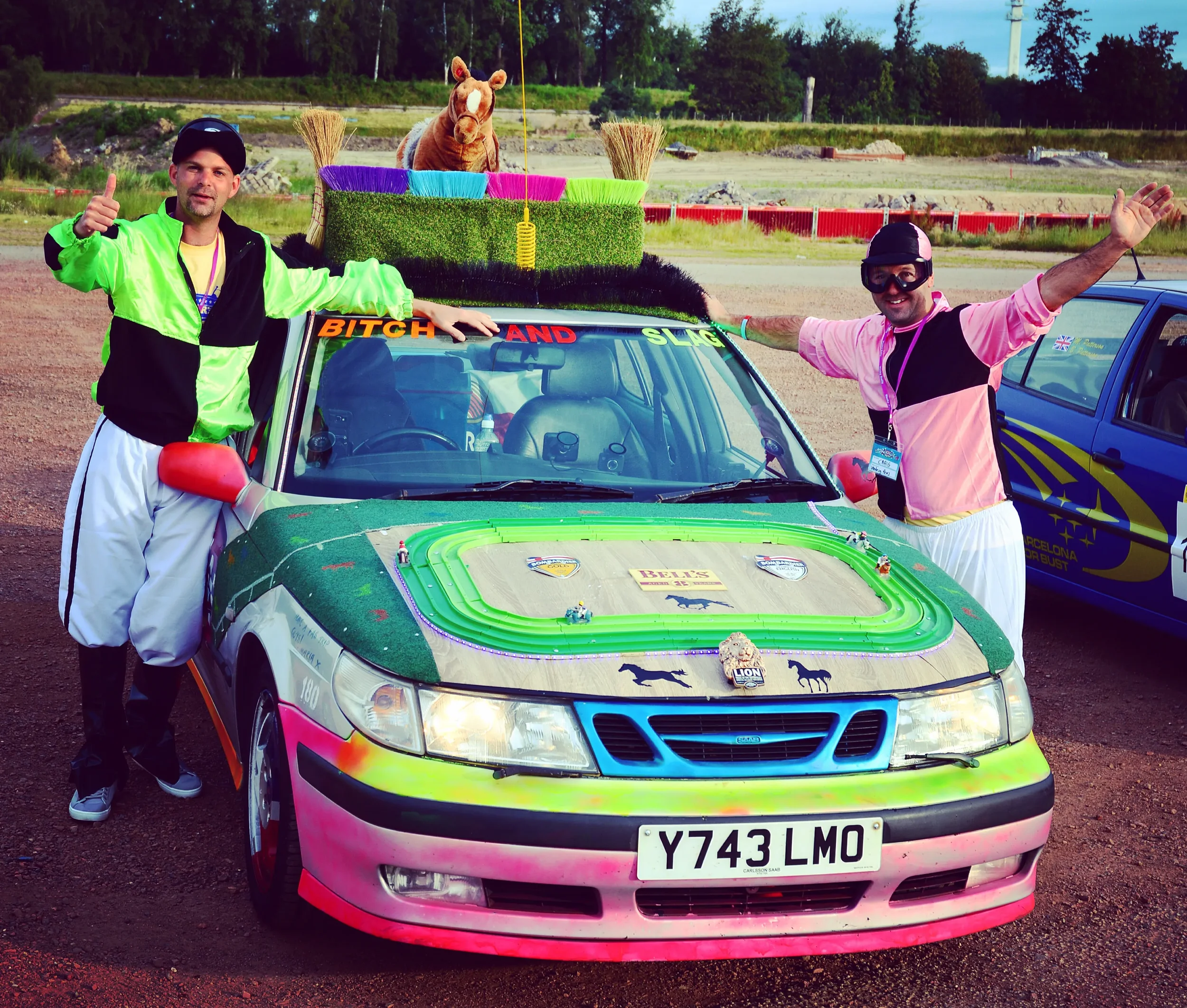 Two men in colorful outfits standing next to a decorated car with a stuffed horse on top. The car has a bright, multicolored paint job and various decorations, including a sign on the windshield that reads 'BITCH AND SLAG.' The men are smiling and po