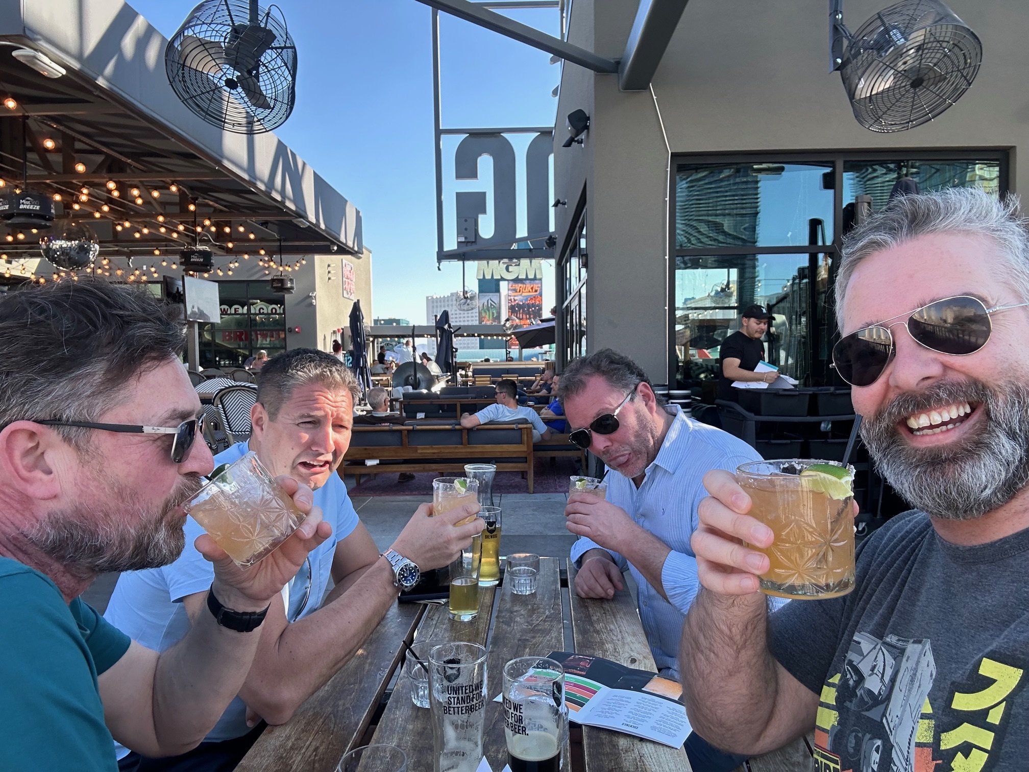 Four men sitting at an outdoor bar or restaurant, smiling and enjoying drinks, with a cityscape background including the MGM sign and a clear blue sky.