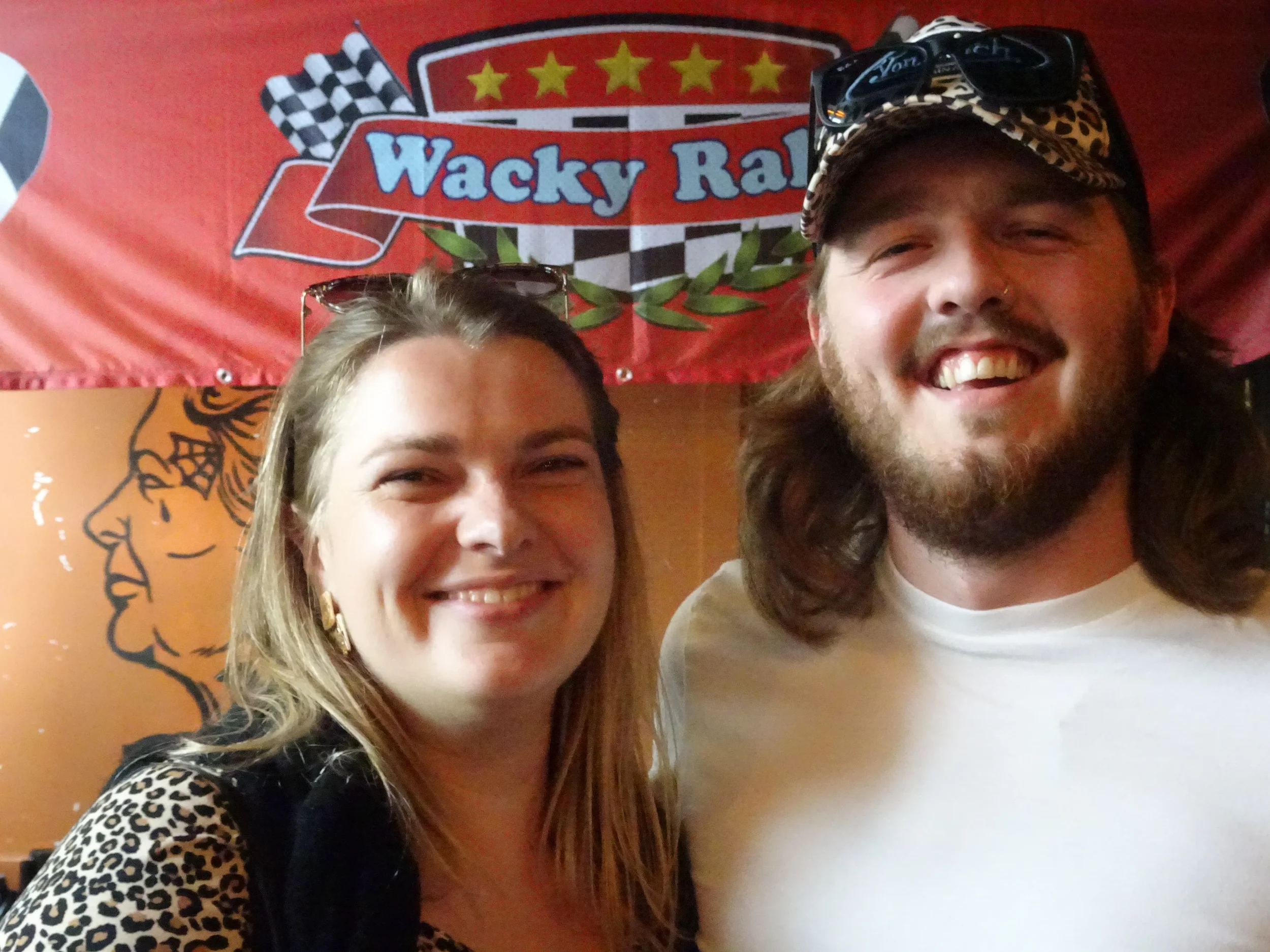 A smiling woman with long blonde hair and a man with long brown hair and a beard, both posing in front of a red banner with a racing-themed logo that reads 'Wacky Races' and checkered flags. The woman is wearing a black top with leopard print sleeves