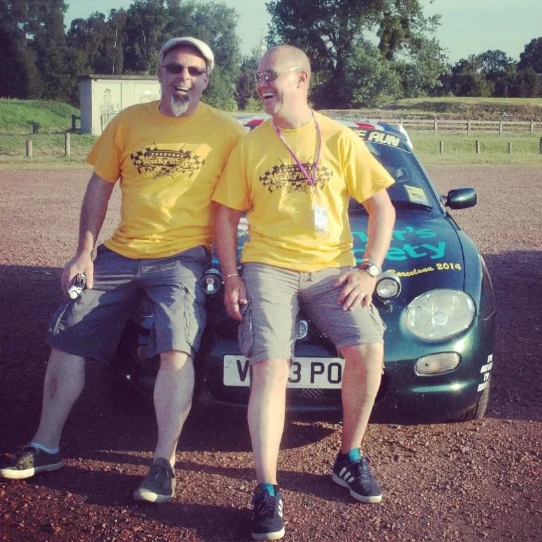 Two men wearing yellow T-shirts sitting on the hood of a green police car, smiling and laughing outdoors during daytime.