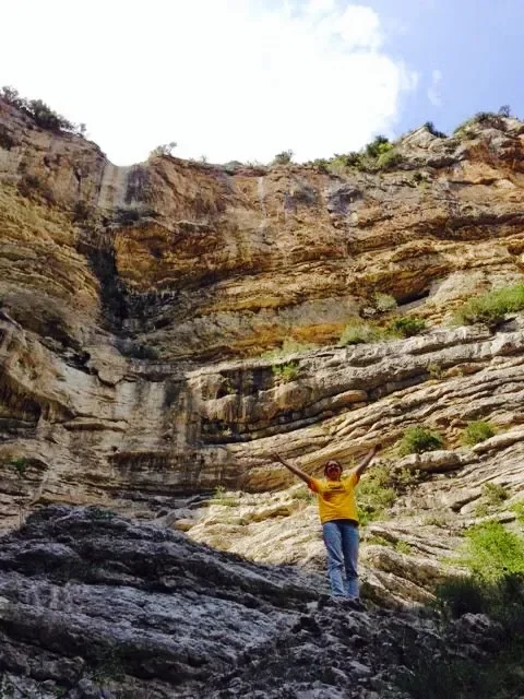A person standing on rocky terrain in front of a steep, layered canyon wall with arms raised.
