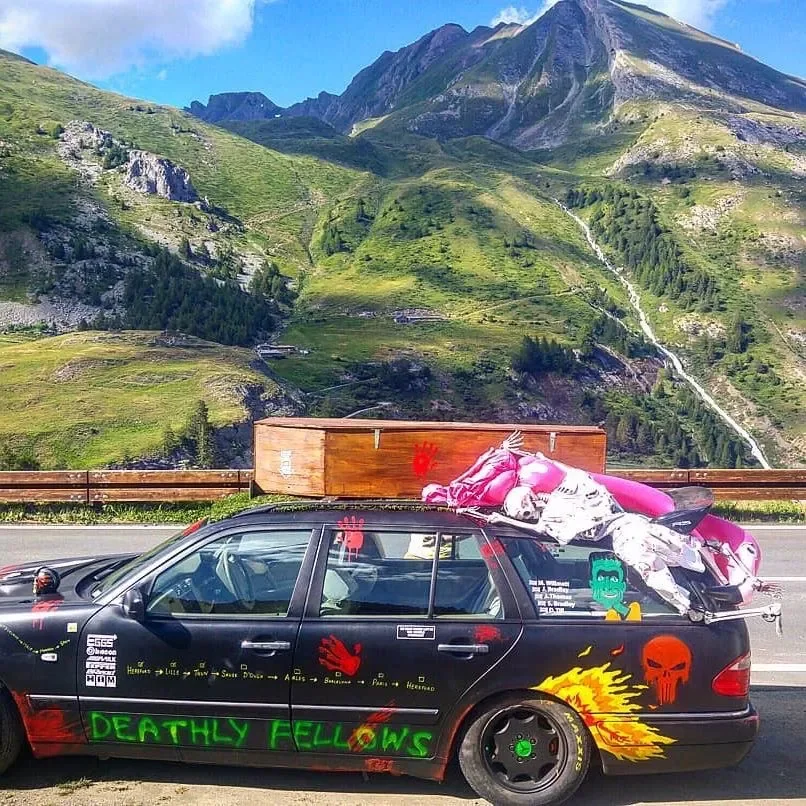 A black car with colorful painted designs, including green text saying 'Deathly Follows,' red handprints, red skulls, and flames; carrying a pink motorcycle on its roof; parked on a mountain highway with green hills and a waterfall in the background.