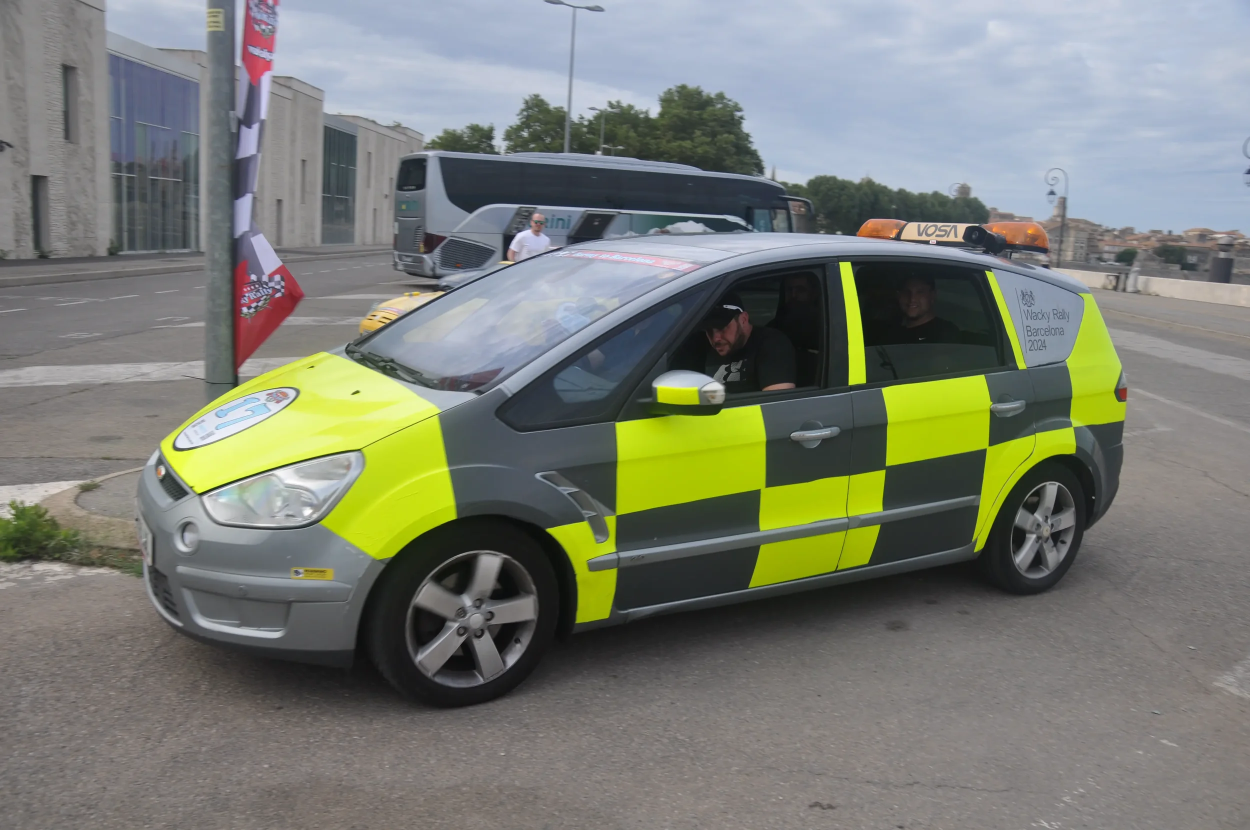 A yellow and gray emergency vehicle with flashing lights parked on a street, with two people inside. In the background, a bus and some pedestrians are visible.