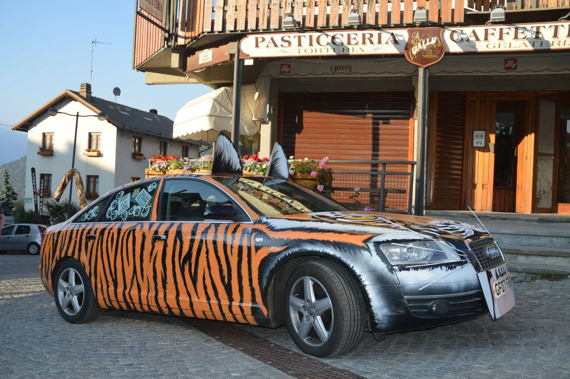 A black Audi car decorated with orange and black tiger stripe patterns, with fake tiger ears attached to the roof, parked in front of a building with a sign that reads "Pasticceria Tortería Gelly." The car has tiger face designs painted on the hood.