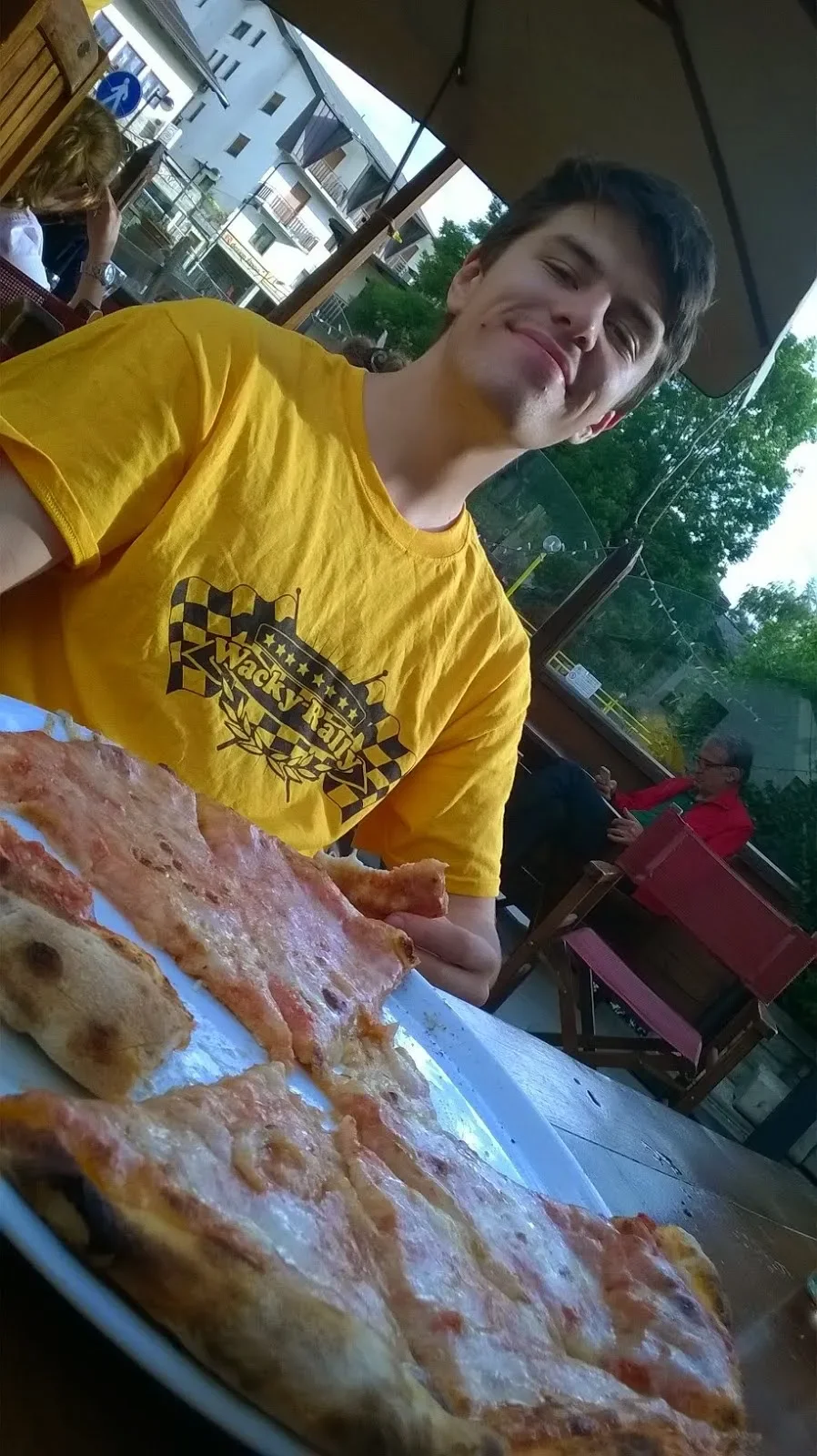 A young man in a yellow t-shirt sitting outdoors at a restaurant table with pizzas in front of him, smiling. In the background, an older woman in a red jacket is seated, with outdoor buildings and trees visible behind her.