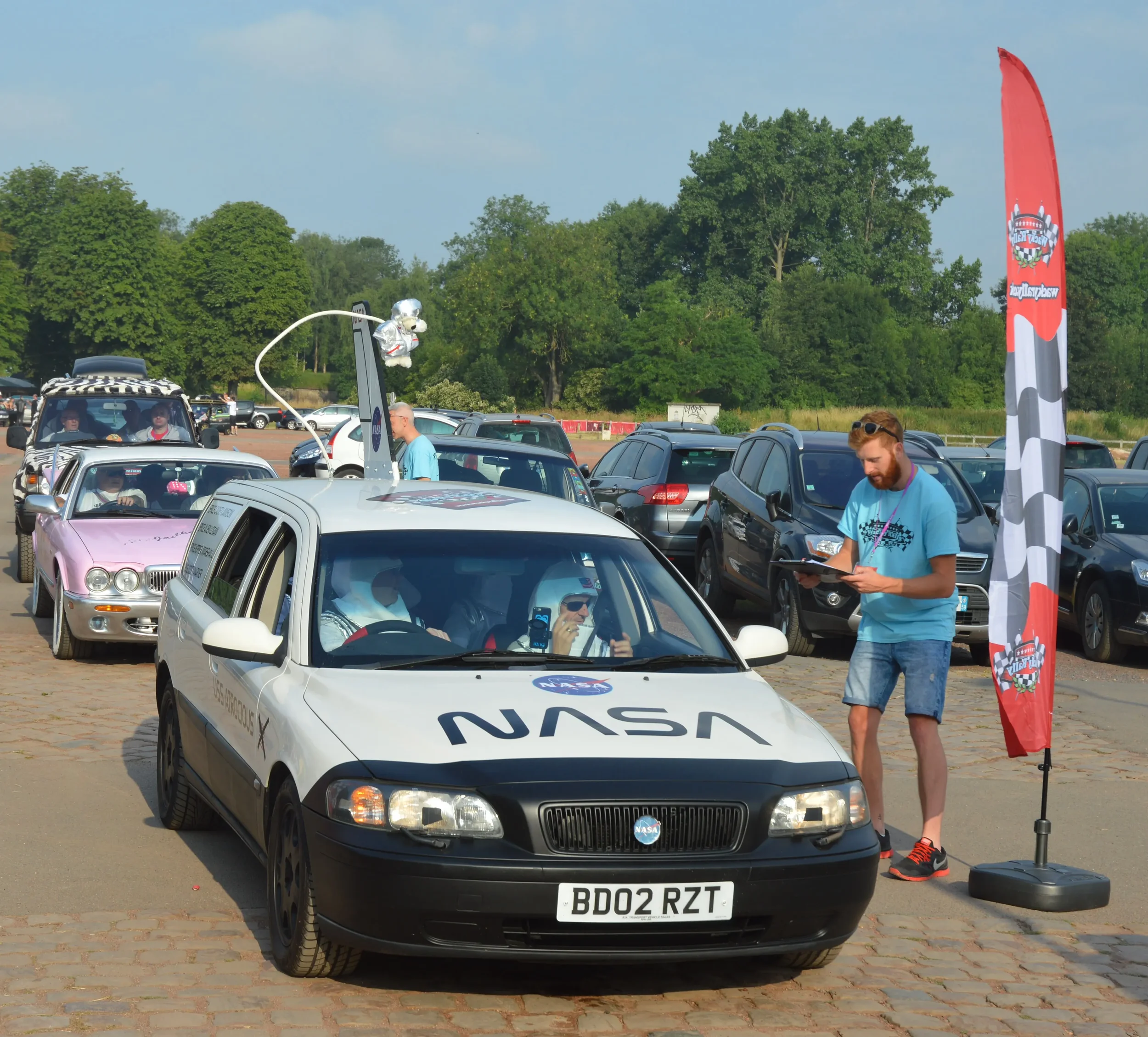A NASA-themed vehicle parked in a parking lot during a car event, with a flag and a person reading a booklet nearby, and other cars and trees in the background.
