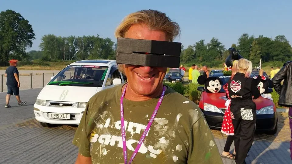 Man with a cardboard VR headset smiling outdoors, with cars and people in the background.