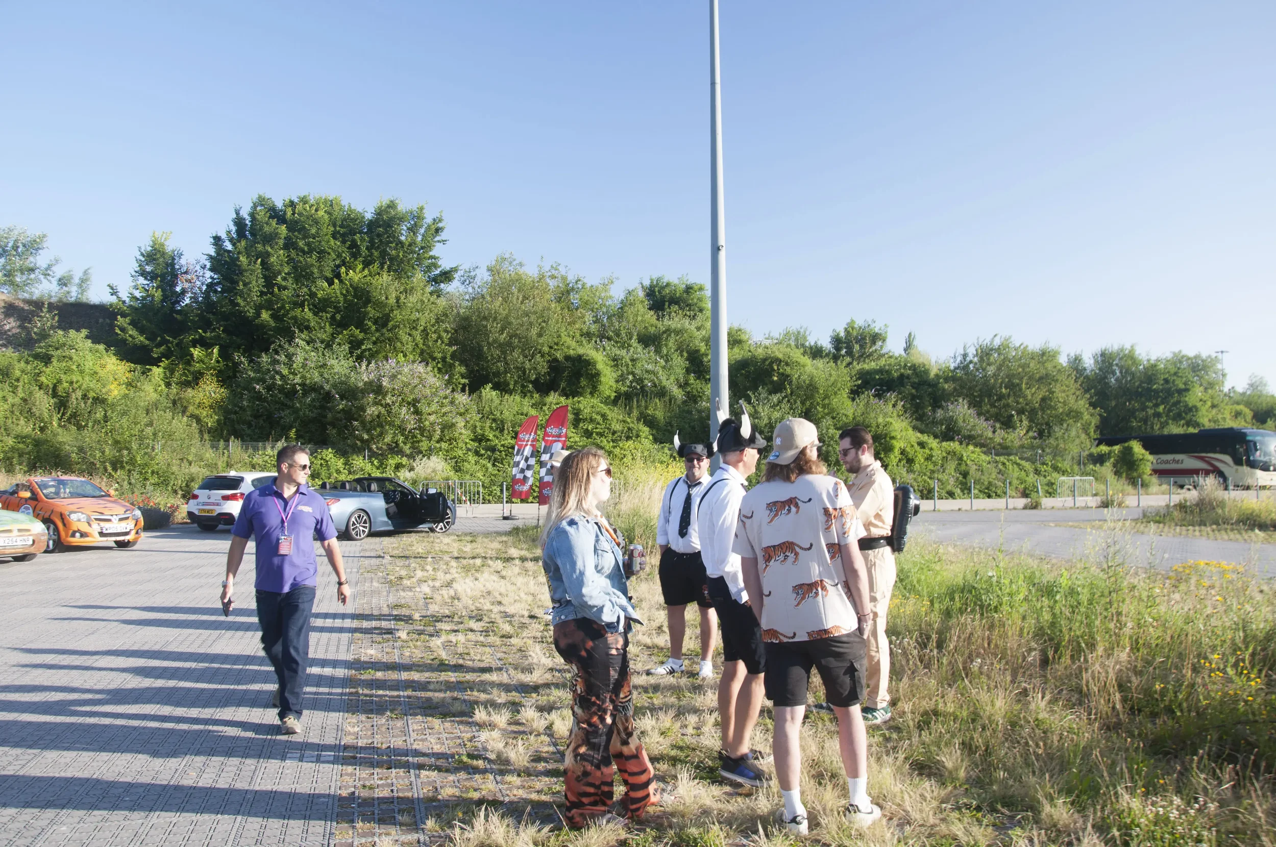 A group of people standing outdoors in a parking lot near green bushes and trees on a sunny day with clear sky, some wearing themed clothing and hats, some holding drinks.