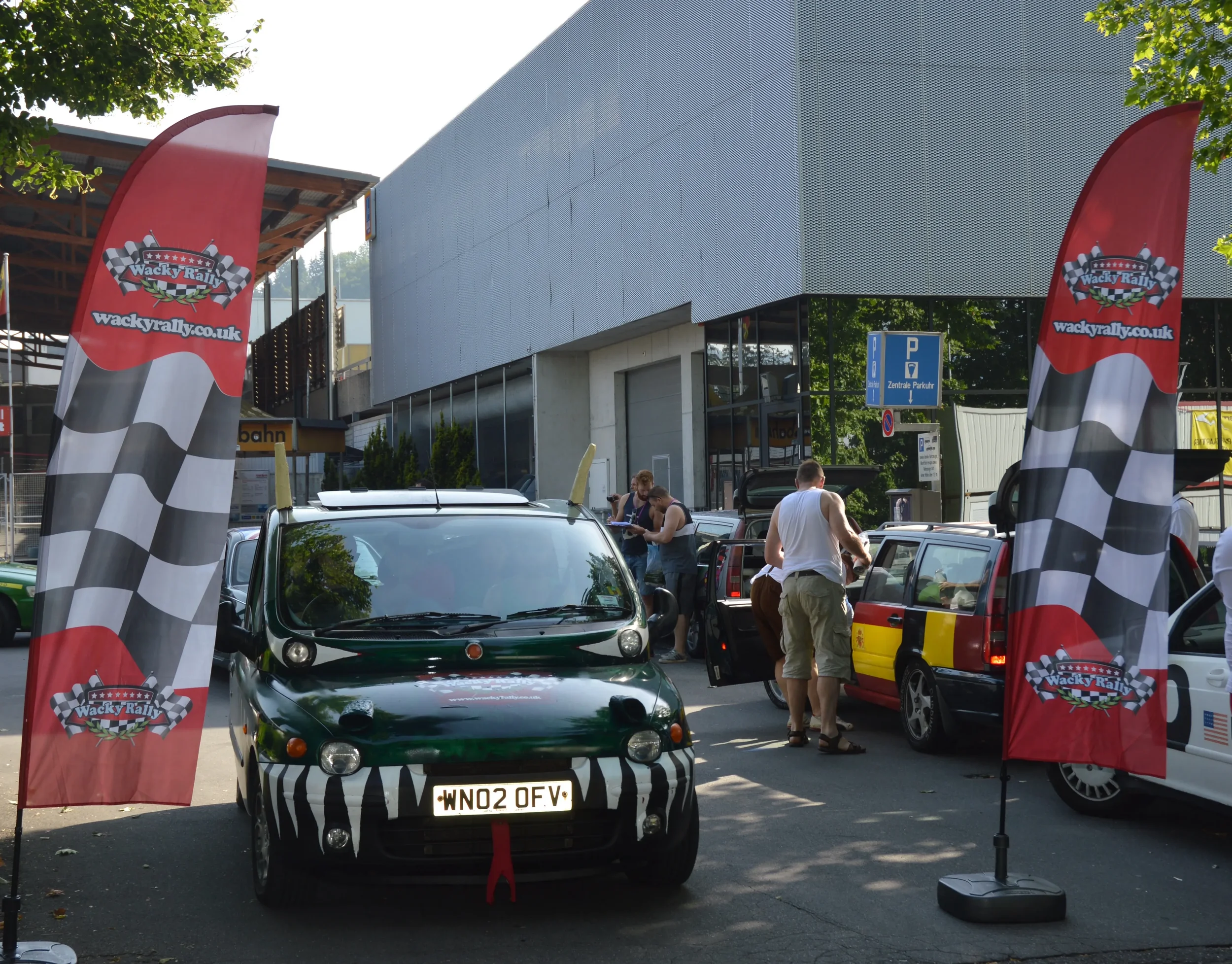 Cars parked at an outdoor event with red banners displaying "Wacky Rally" and checkered flags; people standing around, some looking at cars, in front of a modern building with trees in the background.