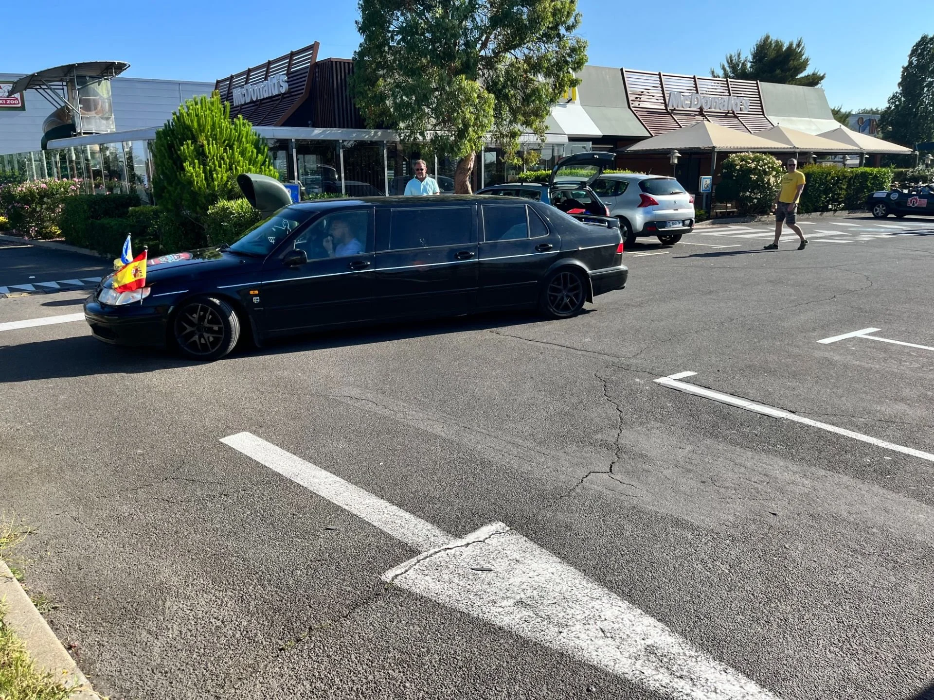 A black car with flags attached to the front parked in a parking lot outside a McDonald's restaurant on a sunny day. There are two men near the car, one inside and one walking nearby, with other cars and people also in the background.
