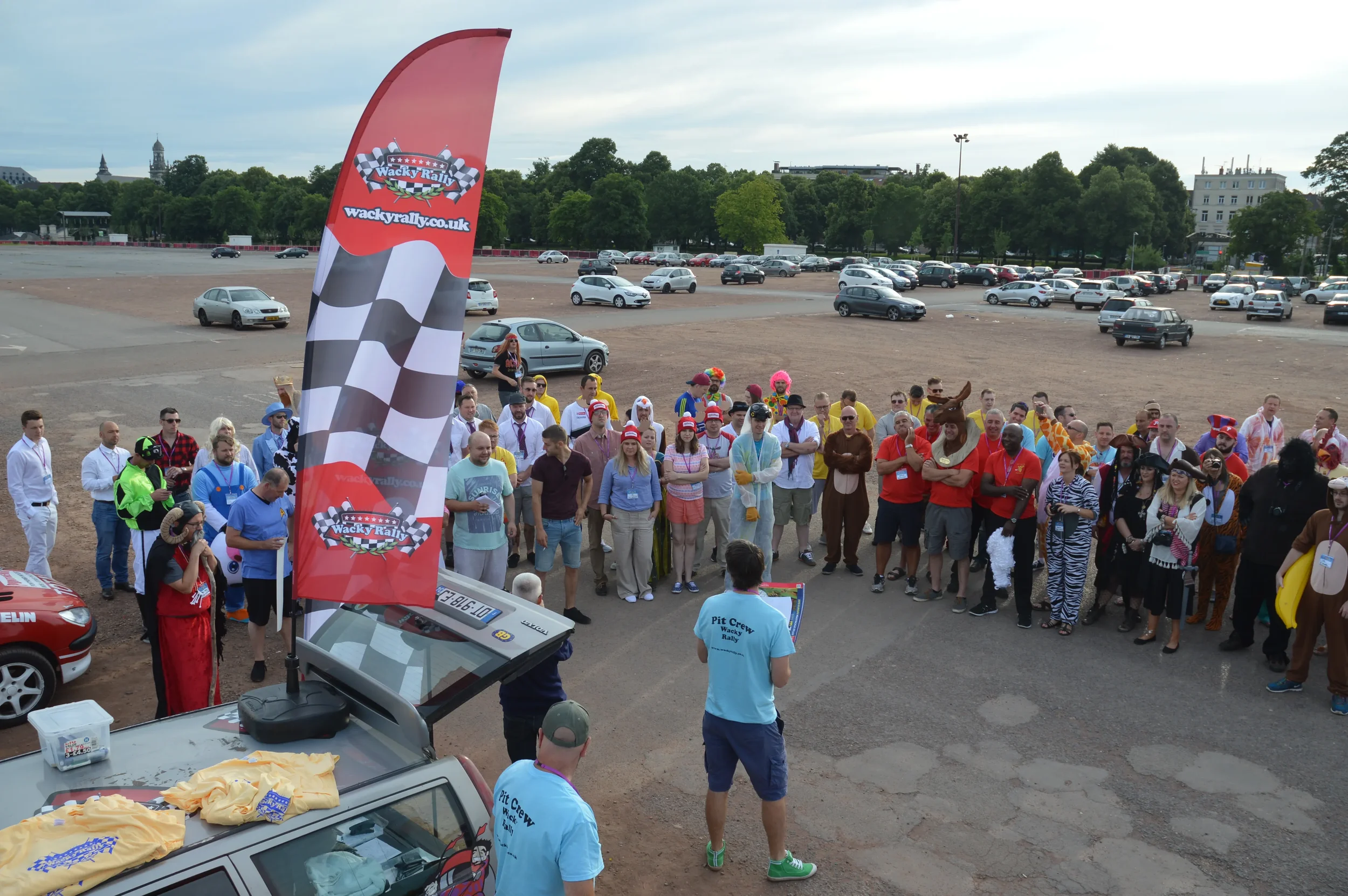 A group of people gathered outdoors in a large open parking lot, many dressed in colorful costumes and casual clothing, listening to a person speaking at a stand with a Wacky Rally flag, during daytime with parked cars and trees in the background.