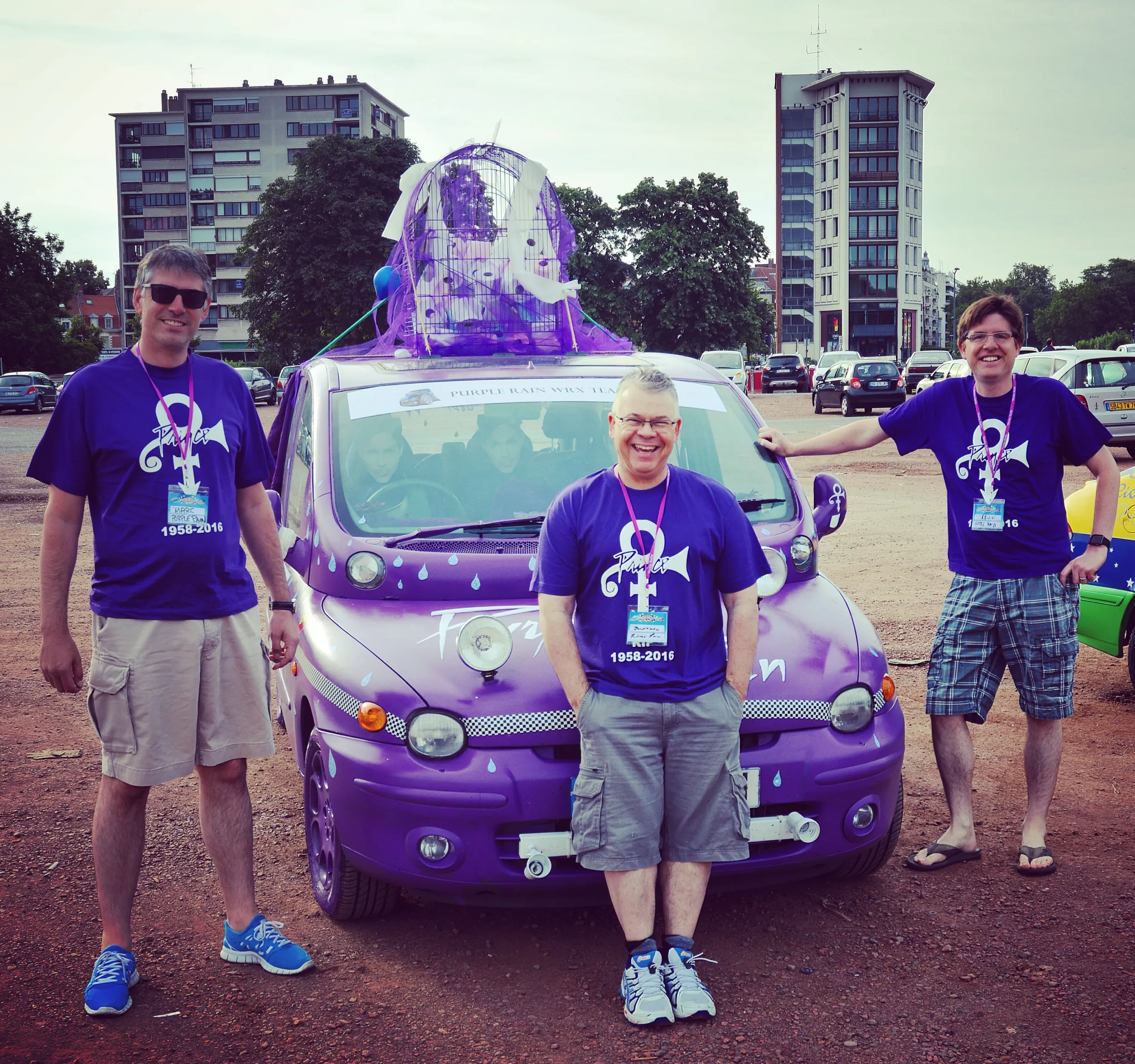 Three people standing beside a decorated purple compact car at an outdoor event. The car has a large purple and white rain cloud decoration with raindrop decals, and the people are wearing matching purple T-shirts with a shark and whale graphic. Two 