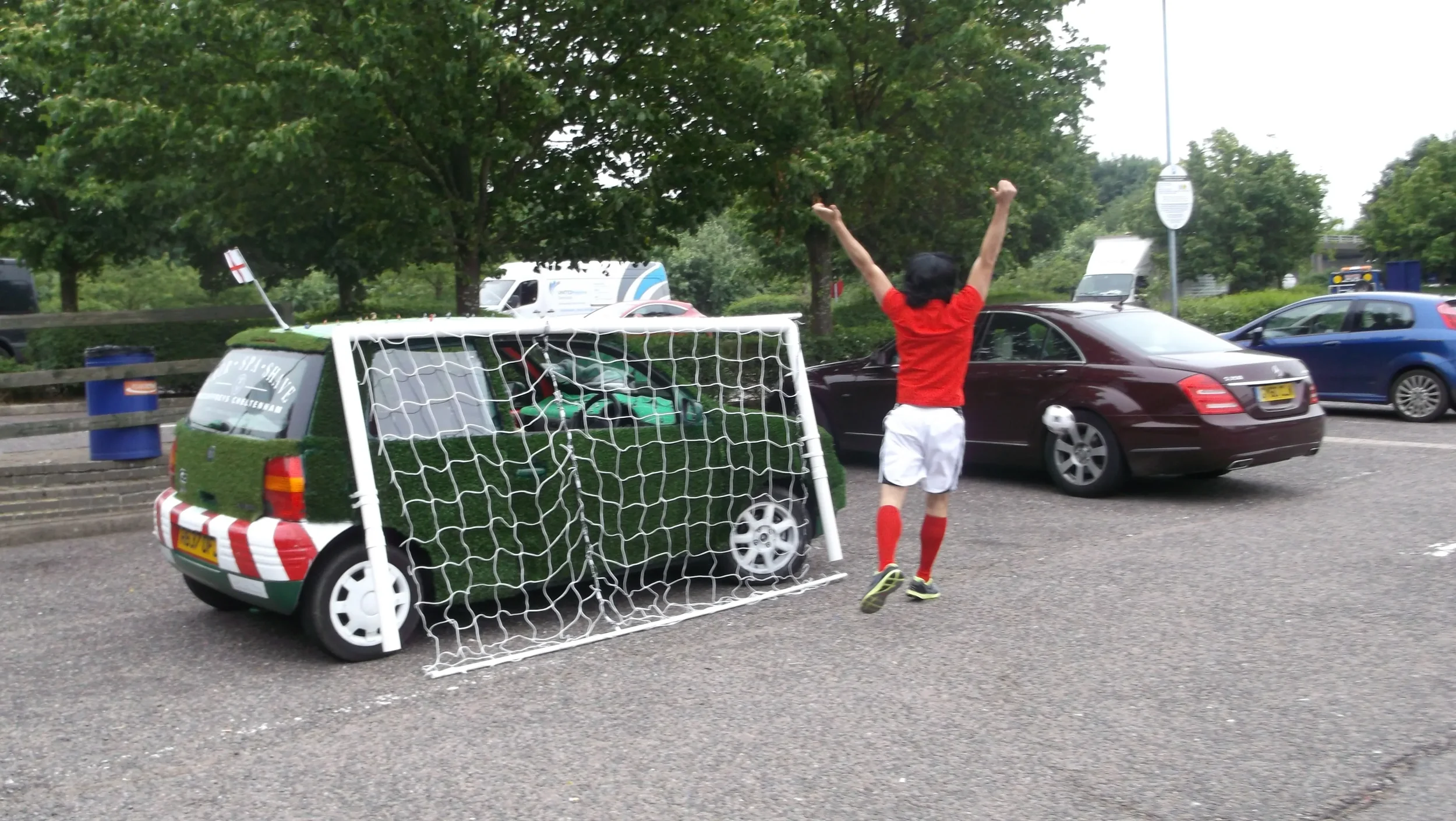 A person in a red shirt, white shorts, and red socks celebrating after hitting a soccer ball into a makeshift goal in a parking lot. The goal is made of small white nets attached to a small car covered in green paint resembling grass. There are trees