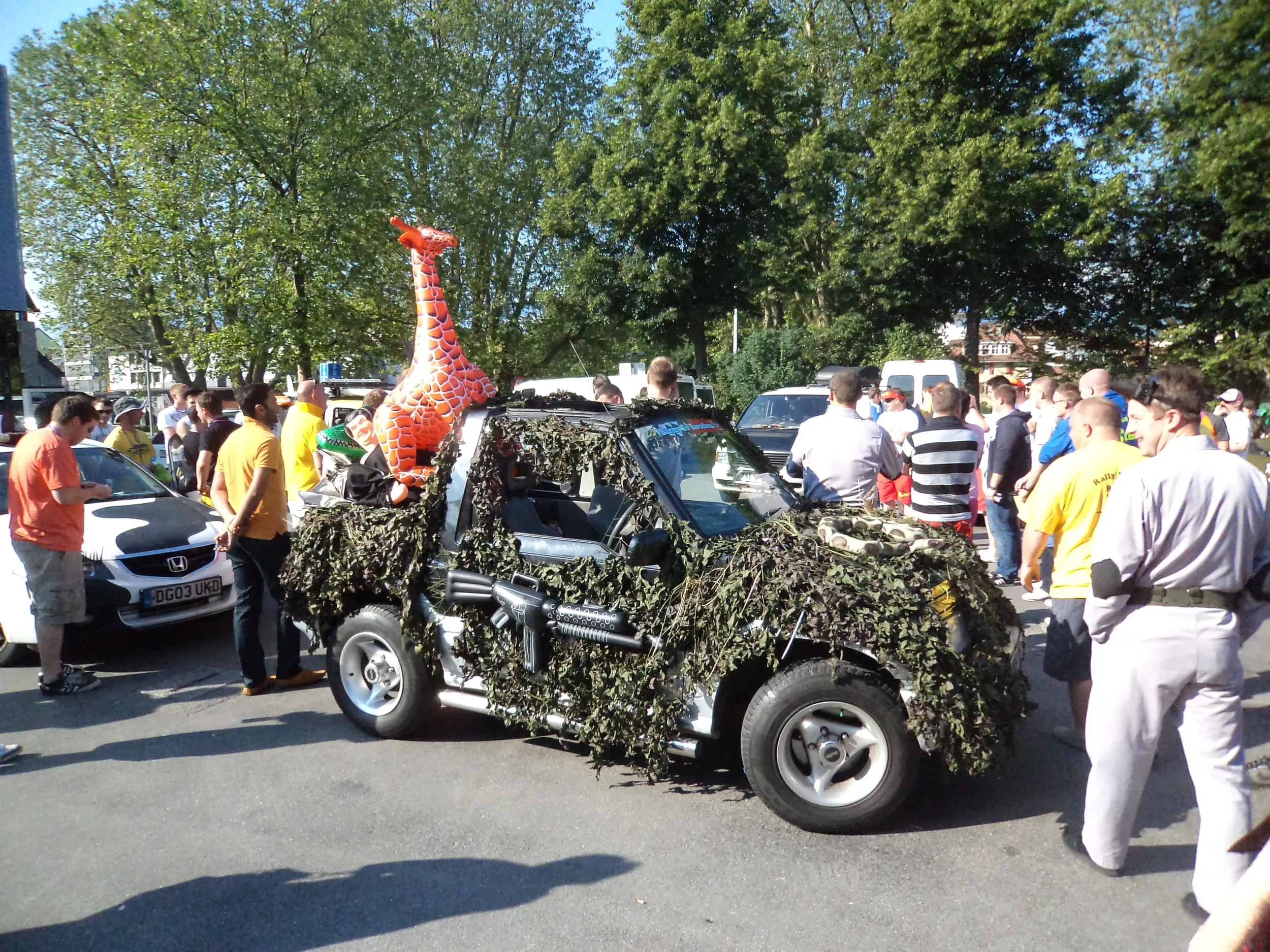 A camouflage-covered vehicle with a large inflatable giraffe attached to it, surrounded by a crowd of people at an outdoor event.