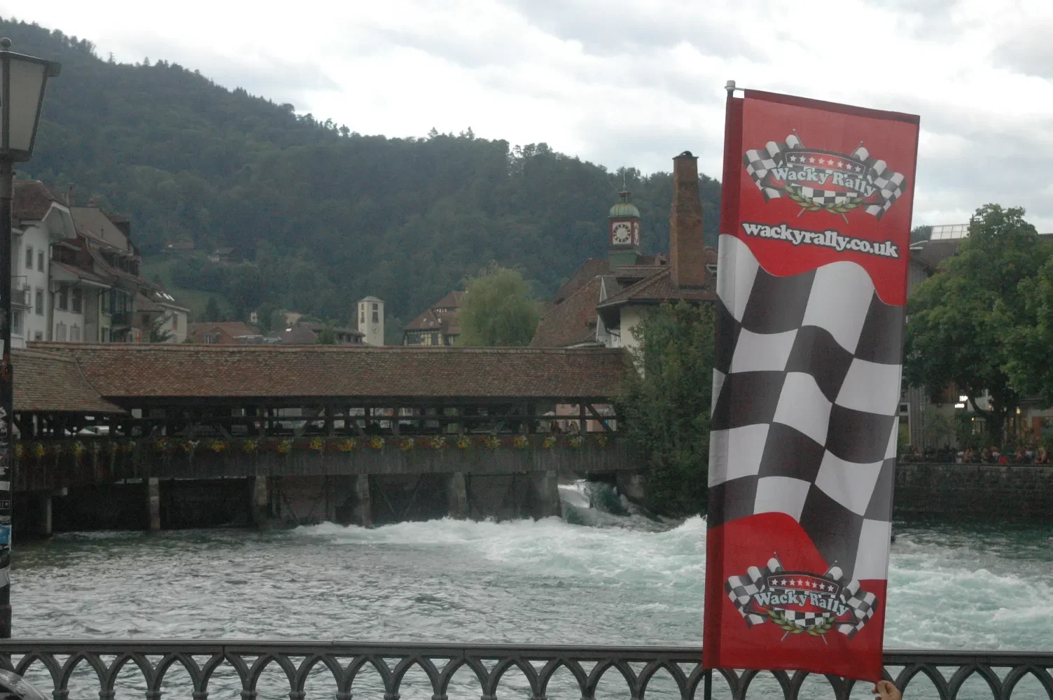 A river with a dam and a covered bridge in a town with buildings, lush green hills, and a cloudy sky in the background. A red banner with a checkered flag and the text 'Wacky Rally' is in the foreground.