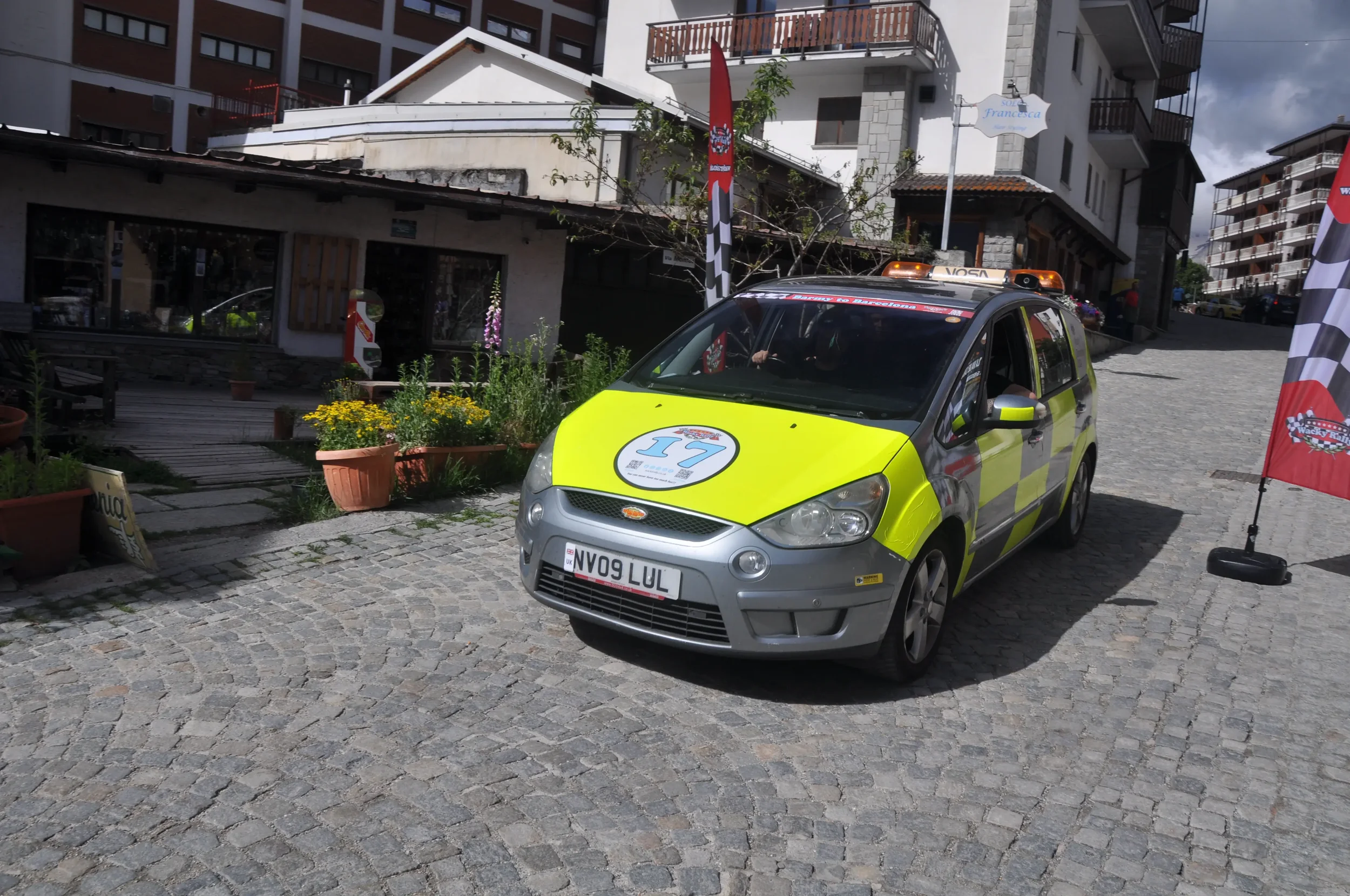 A yellow and gray emergency vehicle with the number 17 on the hood is parked on a cobblestone street with buildings and plants in the background.