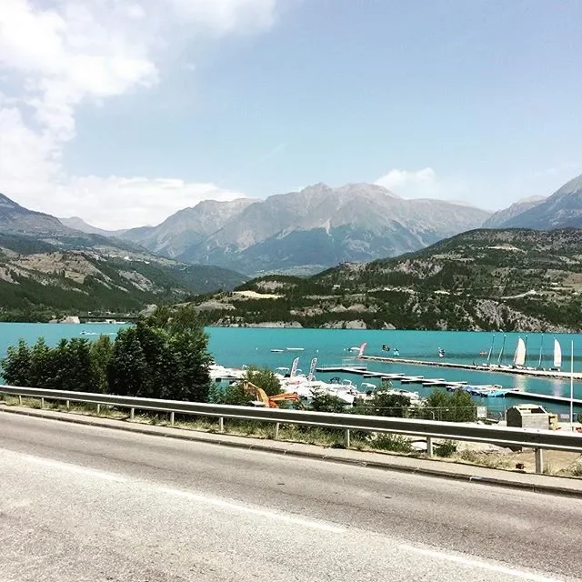 Lake with boats and sailboats, surrounded by green hills and mountains under a partly cloudy sky.