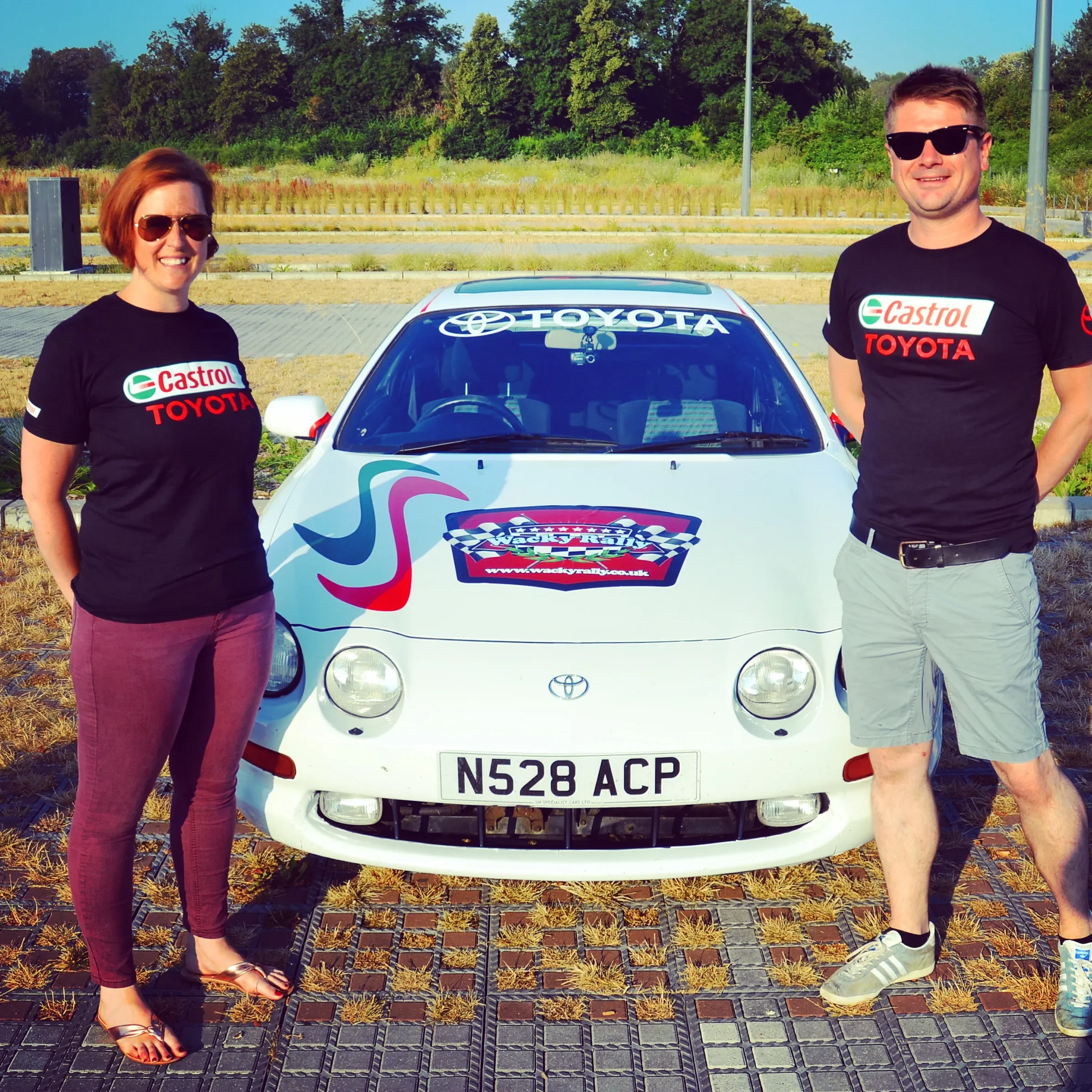 A woman and a man standing next to a white Toyota rally car with racing decals, both wearing black Castrol Toyota t-shirts and sunglasses, in an outdoor parking area with green trees and clear skies.
