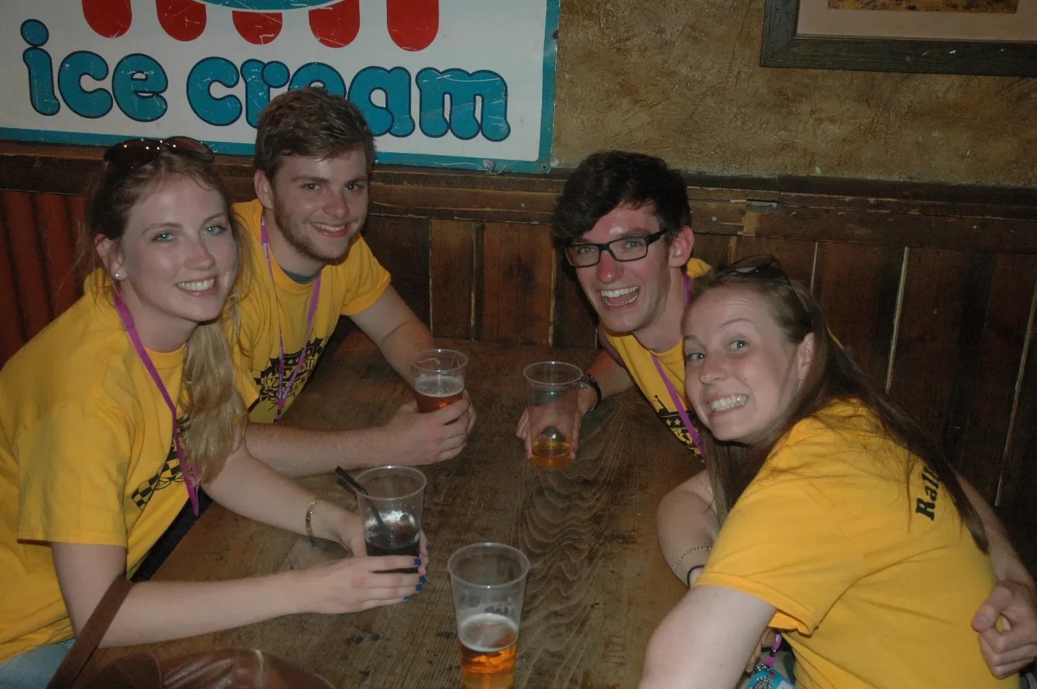 Four young adults in yellow shirts smiling and holding drinks at a wooden table in a bar or pub.