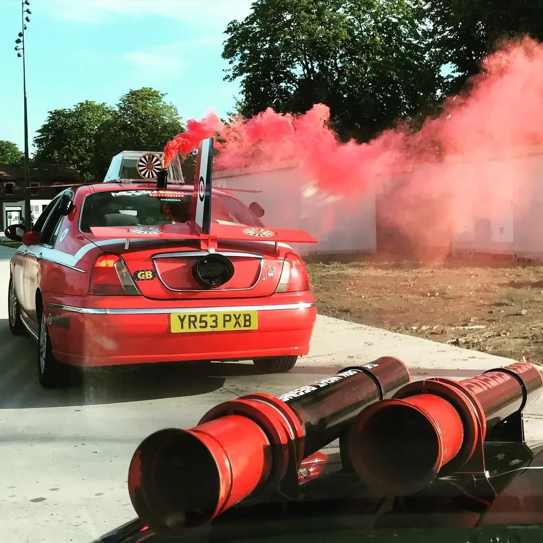 A red car with a racing-themed spoiler and a customs plate YR53 PXB, is on a race track. There are red smoke flares and smoke clouds around, indicating a racing event or celebration.