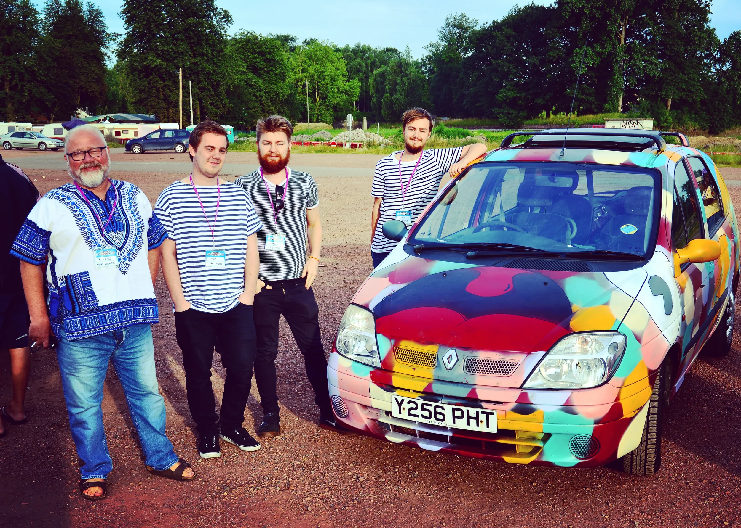 Five men standing next to a colorful, painted Renault car at an outdoor event, with trees and parked cars in the background.