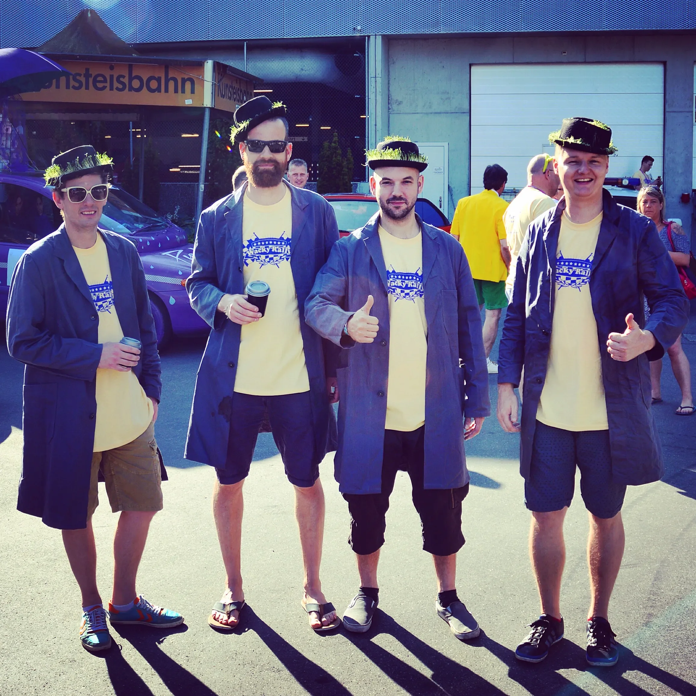 Four men wearing matching yellow shirts and dark blue jackets, with black hats and accessories, standing outdoors, some holding drinks, smiling or giving thumbs-up, in front of a colorful vehicle and a building.